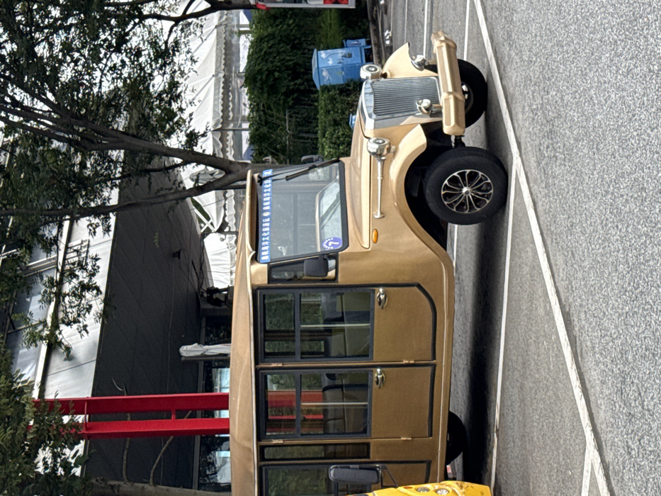 A gold-colored, retro-styled electric tour bus, designed with classic automobile aesthetics, is stationary in a paved parking area within the Shinan District of Qingdao, China. The vehicle exhibits a chrome-like front grille, circular headlights, a gold-colored bumper, and multiple framed glass windows along its side. A blue banner across the windshield identifies it with the 'Qingdao Olympic Sailing Culture Tourism Area,' and a blue circle with the number '7' is affixed to the passenger side windshield. The ground is dark gray pavement marked with white parking lines. In the background, deciduous trees partially obscure a modern building featuring a dark gray facade and prominent red structural columns, as well as a large white event tent. A blue waste receptacle is positioned near green shrubbery. No active human presence is distinctly identifiable.