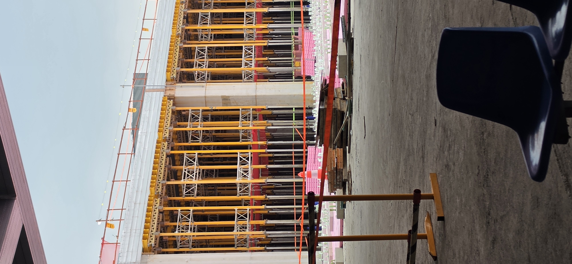The image depicts an outdoor construction site in Pyeongtaek-si, South Korea. In the foreground, two dark blue plastic chairs are visible, one in focus and another slightly out of focus to the right. Behind the chairs, a construction barrier made of orange poles and black and yellow striped tape is present. Further back, a large structure is under construction, characterized by extensive yellow scaffolding and concrete pillars. The ground is paved with concrete or asphalt. A single orange and white traffic cone is also visible in the middle ground. The sky above is a pale, overcast blue. The overall scene suggests an active but paused construction project.
