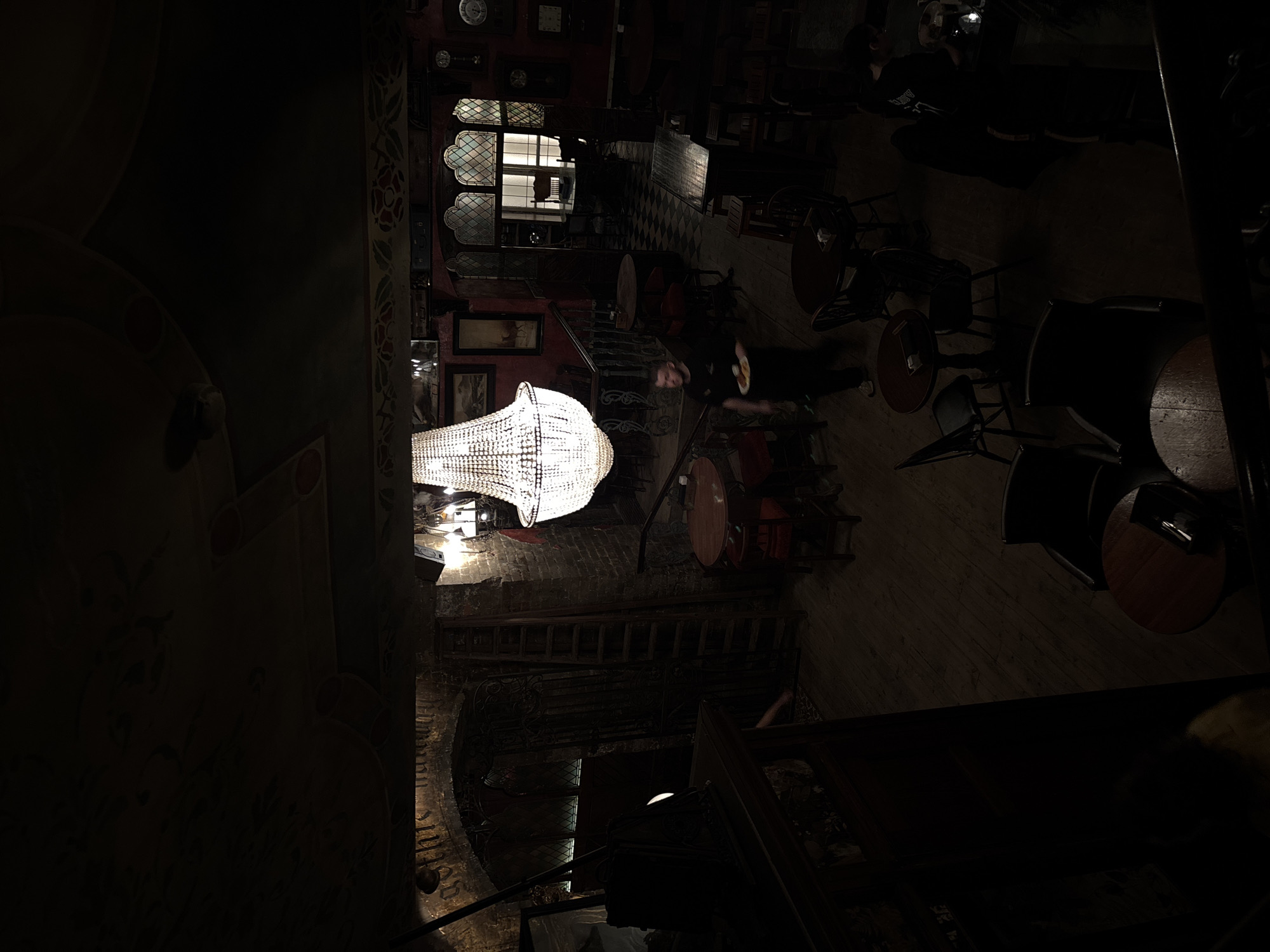 The image shows an interior view of a dimly lit, multi-level space that seems to be a restaurant or bar. The location is the Didu restaurant in Moscow, Russia.
A large, ornate chandelier hangs prominently, providing most of the visible light. Tables and chairs are scattered around the wooden floor, some of which are occupied.
A person is standing at the top of the stairs, who looks to be a waiter wearing dark clothes. A patterned wall features windows with lattice designs. There is a flight of stairs leads to a lower level, where there is writing on the wall. There are also ladders next to the stairs that look like they have been fixed for design.