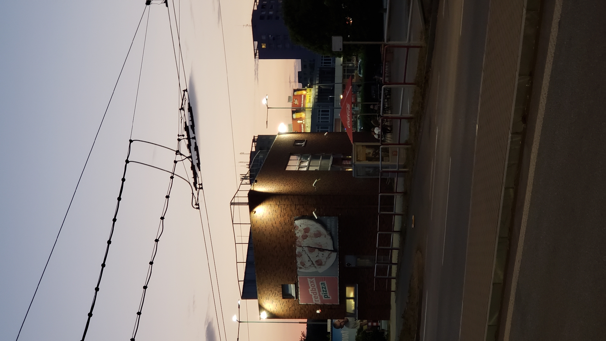 A brick building with a flat roof and a railing around it is illuminated by exterior lights. A large sign on the side of the building advertises "redbox pizza" and shows a picture of a pizza. There are windows on the building, some with blinds. In front of the building, there is a sidewalk with a red and white fence and a bus stop sign. A few people are seated at tables under a red umbrella outside the building. Across the street, there is a road and several other buildings, including a tall, dark apartment building and buildings with illuminated billboards. The sky is a pale gradient of purple and orange, indicating dusk or dawn, and overhead, trolley wires are visible against the sky.