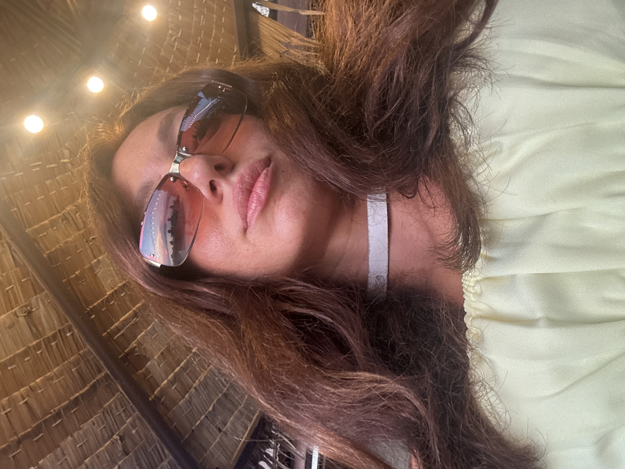 A close-up, low-angle portrait features a woman with long, dark brown hair, possibly taking a selfie, set against a backdrop of a woven ceiling. Her head is slightly tilted, and her lips are softly parted, suggesting a relaxed or pensive expression. She wears large, reflective sunglasses that obscure her eyes; the lenses vividly reflect a cloudy sky, hinting at either sunrise or sunset with warm hues of orange, pink, and blue, and possibly distant structures.
She is wearing a light-colored top, likely pale yellow or green, with gathered or ruched fabric visible on her right side. The background reveals a distinctive ceiling constructed from tightly woven straw or bamboo mats, characteristic of traditional Thai architecture or resort buildings. Three bright, circular artificial lights are embedded in the ceiling above her. The overall ambiance, consistent with the location context of Baan Plai Laem, Thailand, suggests a serene, tropical setting, likely indoors or under a covered outdoor area. There is no visible text in the image.