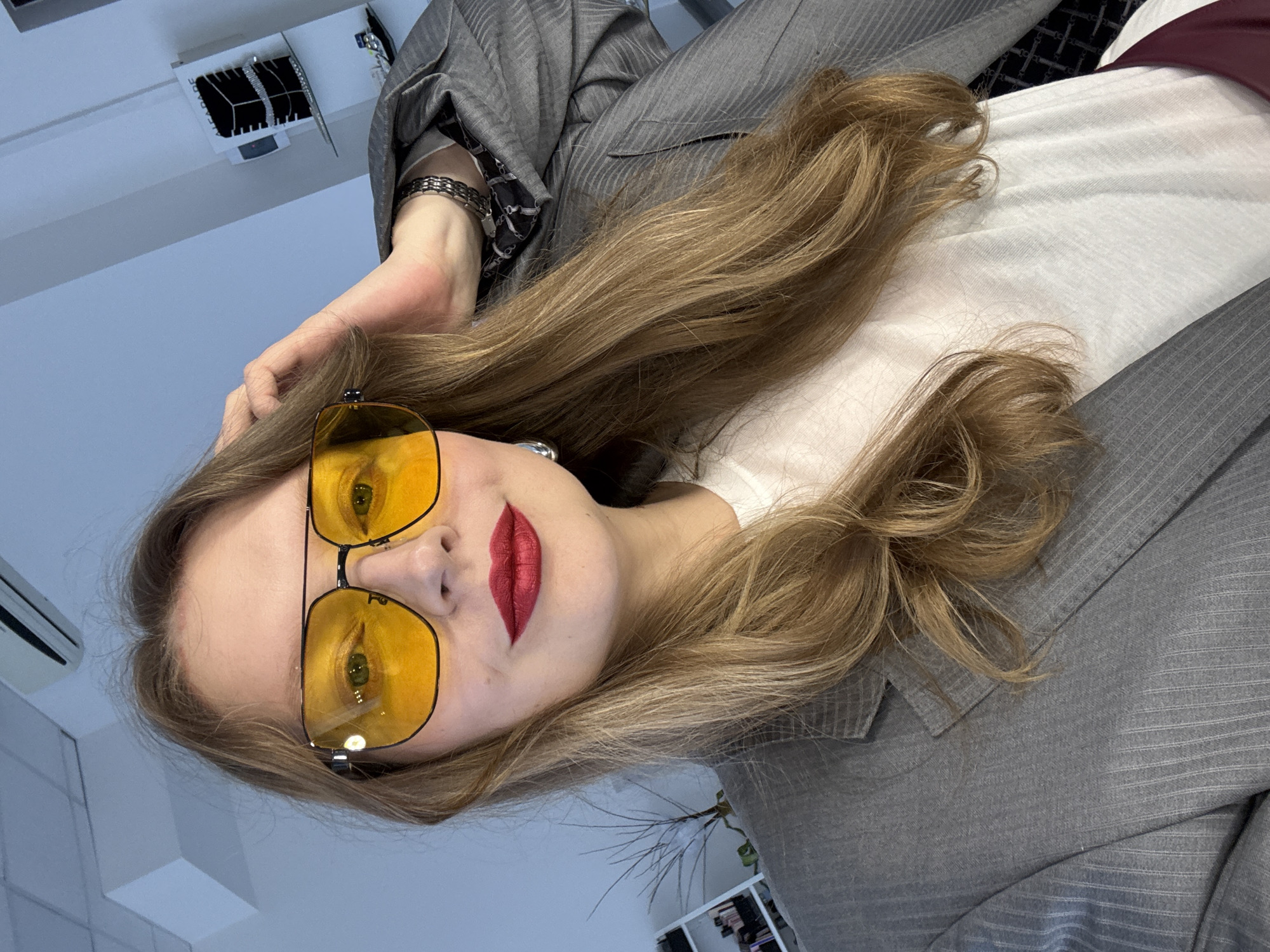 An adult female is depicted in a close-up selfie, looking directly at the camera. She has long, wavy, light brown hair styled to fall over her shoulders. Her face features full lips adorned with vibrant red lipstick, and she wears oversized aviator-style sunglasses with yellow-tinted lenses and dark frames, obscuring her eyes. A silver-toned hoop earring is visible on her left ear. She is wearing a grey pinstripe blazer over a white crew-neck t-shirt. On her right wrist, a multi-link silver-toned bracelet is visible, and the cuff of a dark, patterned garment or scarf can be seen under the blazer sleeve. A dark red or burgundy strap from another garment is visible near the bottom right of the frame. Her right hand is raised, with fingers touching the hair on the right side of her head. The setting is indoors, with light blue-grey walls and a white ceiling from which an air conditioning unit is partially visible in the upper left. In the background on the right, a white wall-mounted display unit features the text "ALFADARE" and holds multiple items, possibly jewelry, presented on dark stands. Below this, a small shelf holds additional objects. On the left side of the background, a white shelving unit is partially visible behind a sparsely branched plant. The scene is located in the city of Izhevsk, Russia.