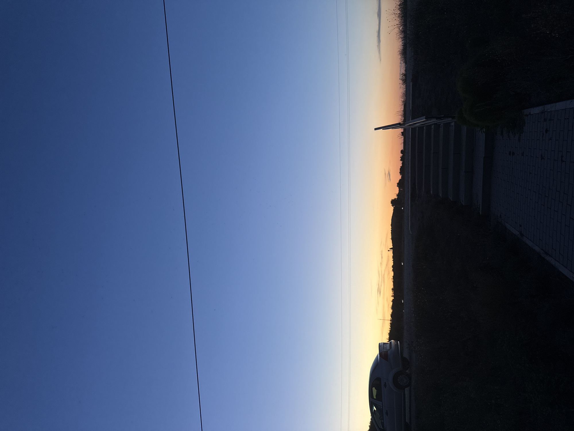 The image captures a twilight scene with a gradient sky transitioning from deep blue at the top to orange and yellow hues near the horizon. A thin, dark horizontal line, likely a power line, stretches across the upper portion of the frame. On the left, a silver sedan is parked on an elevated surface. In the foreground, a paved walkway leads to a set of concrete stairs with a wooden handrail. The area is characterized by dark, silhouetted vegetation and a low-lying landscape with distant lights and structures, possibly indicating an urban or semi-urban periphery. The specific location in Cimișlia, Moldova, is not explicitly discernible from visual cues alone, but the elements suggest an outdoor setting during dusk.