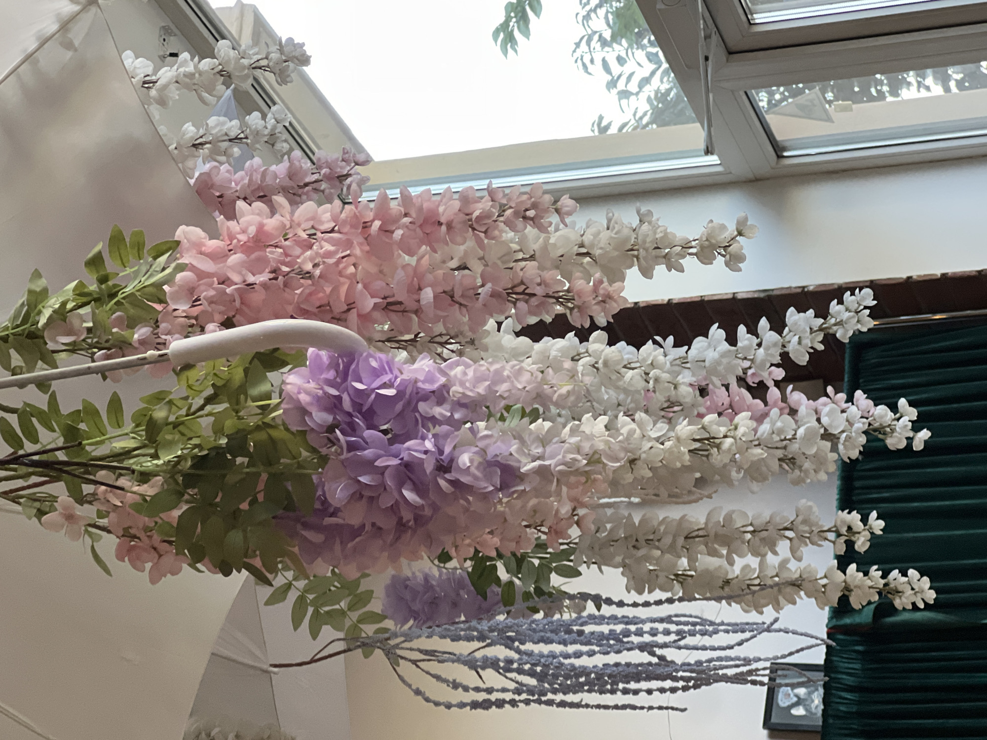A close-up, eye-level shot focuses on hanging artificial wisteria flowers in pastel shades of pink, purple, and white, suspended from what appears to be an open white umbrella. The flowers are arranged in cascading clusters, with green artificial leaves interspersed. To the left, a dark green pleated fabric drapes across the bottom of the frame. Behind this, a framed picture with a dark border is visible. To the right, a large window with white frames allows natural light to enter, revealing outdoor foliage. The scene is indoors, with a white ceiling and a textured brick wall visible on the left. There are no people present. The location is specified as Fontanka, Ukraine.