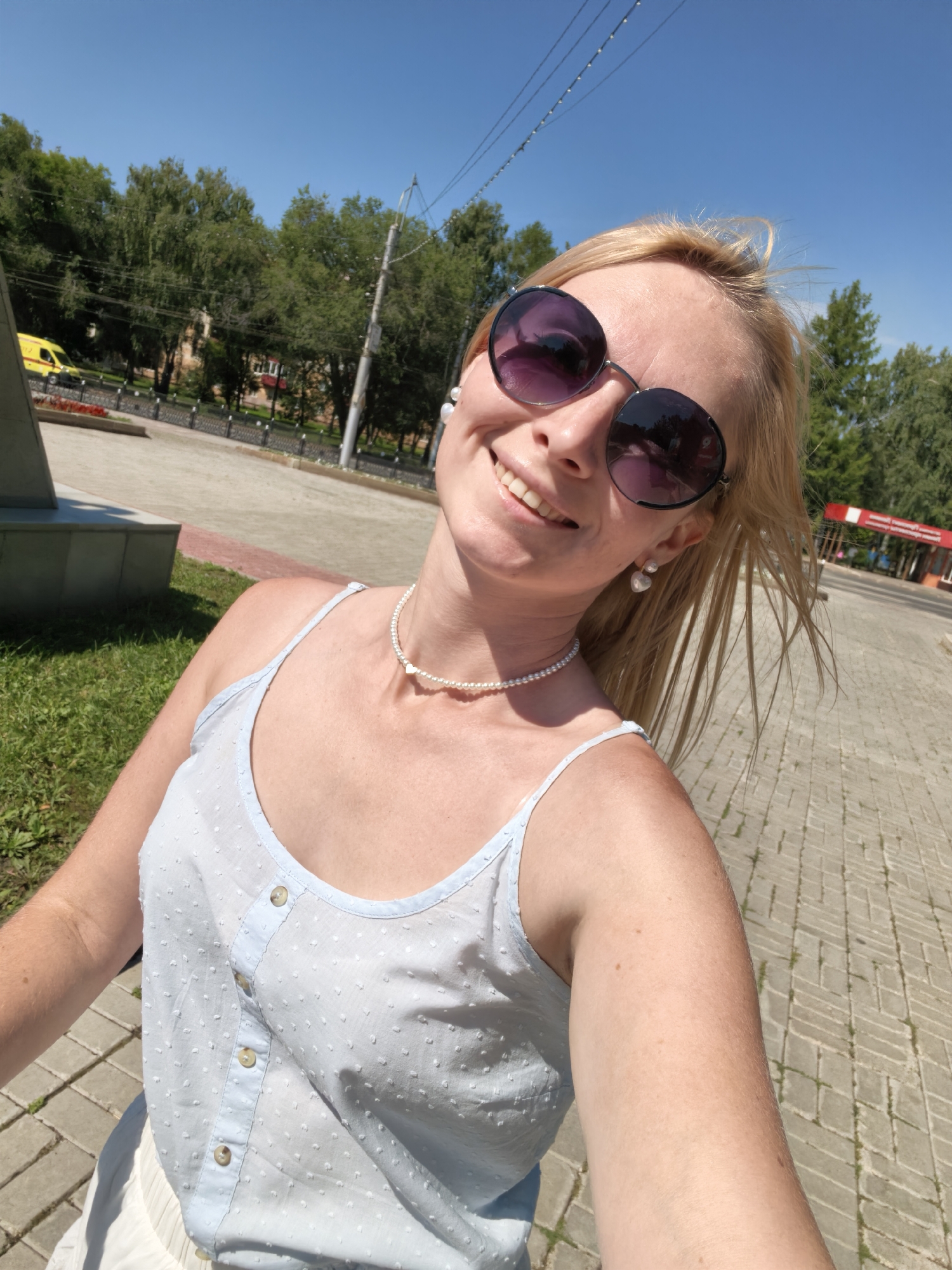 A blonde woman with sunglasses smiles at the camera outdoors. She is wearing a light blue polka dot top, a pearl necklace with a heart pendant, and heart-shaped pearl earrings. The background shows a paved walkway, green grass, trees, and buildings under a clear blue sky. The scene appears to be in the city of Sterlitamak, Russia, based on the provided context.