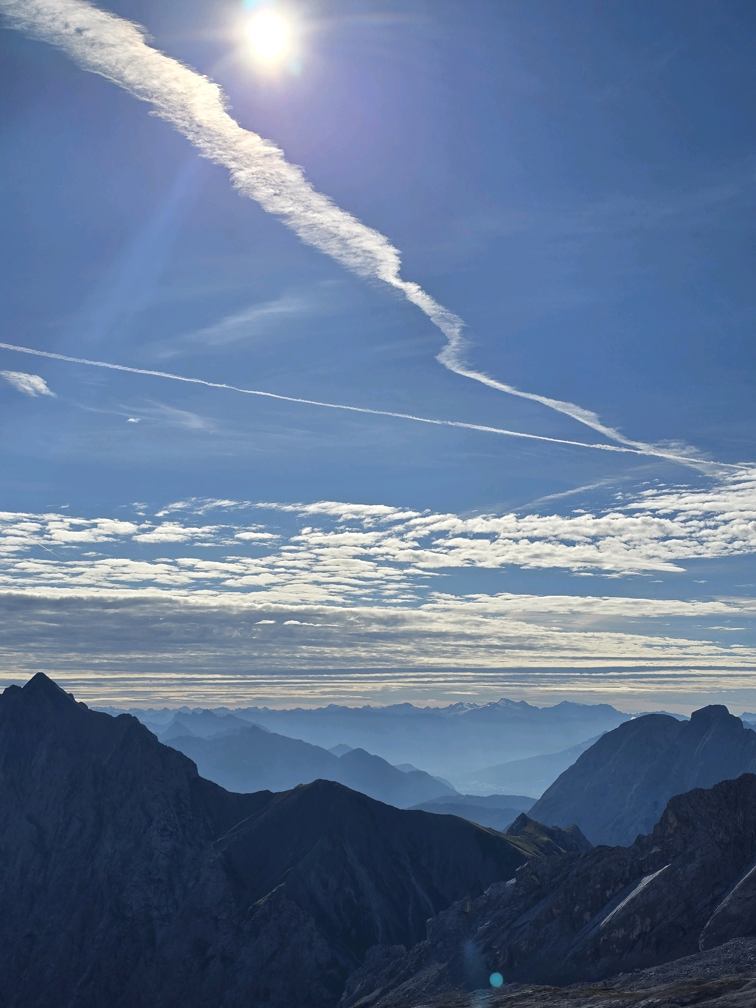 This image captures a panoramic mountain landscape under a vast, blue sky with scattered clouds. The foreground features rugged, dark-hued mountain peaks, suggesting a rocky, alpine terrain. In the midground, successive layers of mountains recede into the distance, becoming progressively lighter and bluer due to atmospheric perspective. The sky is dominated by the bright sun, partially obscured by a wispy contrail stretching across the upper portion of the frame. Below the sun, a delicate band of white, striated clouds runs horizontally, with more layered clouds beneath them. The overall impression is one of a clear, crisp day in a high mountain environment. The location is the Zugspitze area, near Garmisch-Partenkirchen, Germany, renowned for its dramatic alpine scenery. There are no people or animals visible in the image.