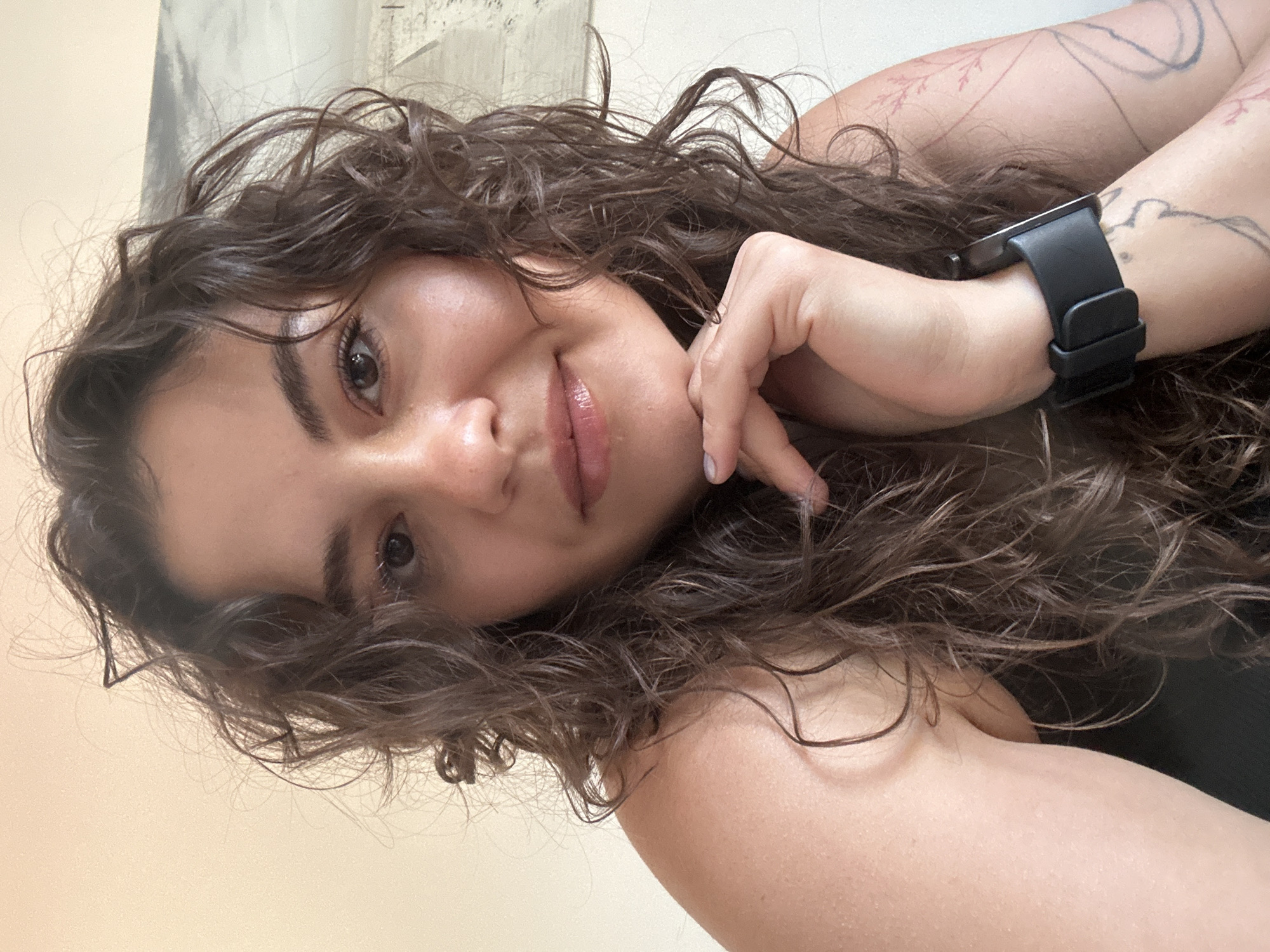 A close-up portrait shows a young woman with curly brown hair lying down. She is fair-skinned and wears a black smart watch on her left wrist. She has tattoos on her left arm. Her hand is resting near her face. The background is a beige wall. The image doesn't provide information about the location in Budva, Montenegro.