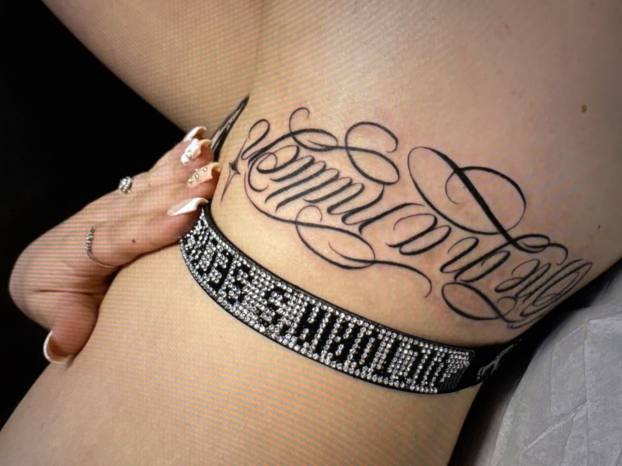 A close-up, overhead shot shows a person's inner thigh with a tattoo and a jeweled garter. The tattoo is in script and reads "Everything". The garter is black with a band of clear rhinestones. A hand with manicured nails is resting on the thigh, with rings on two fingers. The background is black. The image is undated and from an unknown location.