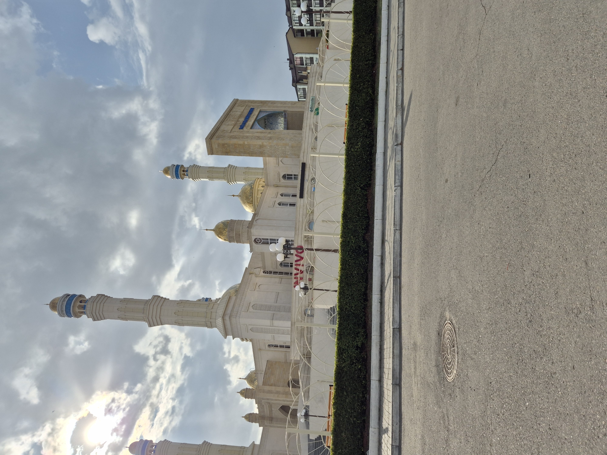 The image shows a mosque in Kokshetau, Kazakhstan, under a cloudy sky with the sun breaking through. The mosque features multiple tall minarets with golden domes and intricate architectural details. A beige-colored facade with arched windows and decorative carvings is visible. In front of the mosque, there is a white decorative fence and a well-maintained green hedge. The foreground displays an asphalt road with a manhole cover and a sidewalk. In the background, several apartment buildings are visible. The overall scene depicts a religious building and its surroundings in an urban environment.