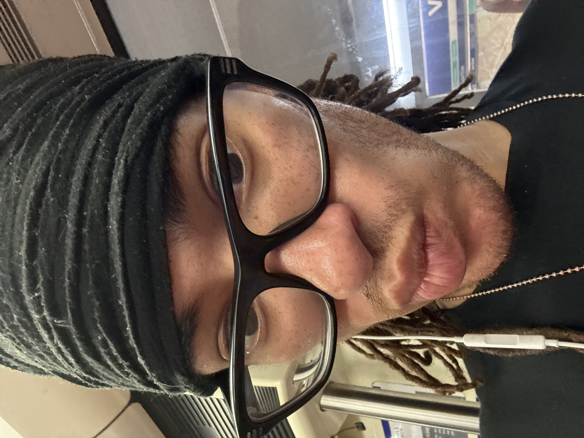 A close-up, angled shot depicts a person with brown skin, wearing black-rimmed glasses and a black head covering with a corrugated texture. The person also has dreadlocks that are visible near their ear. A black shirt and a delicate silver-colored chain are visible at the right of the frame. White headphone wires are visible in the lower right area. The location is likely a train in the city of Paris, France, as indicated by signage visible through the glasses lens.