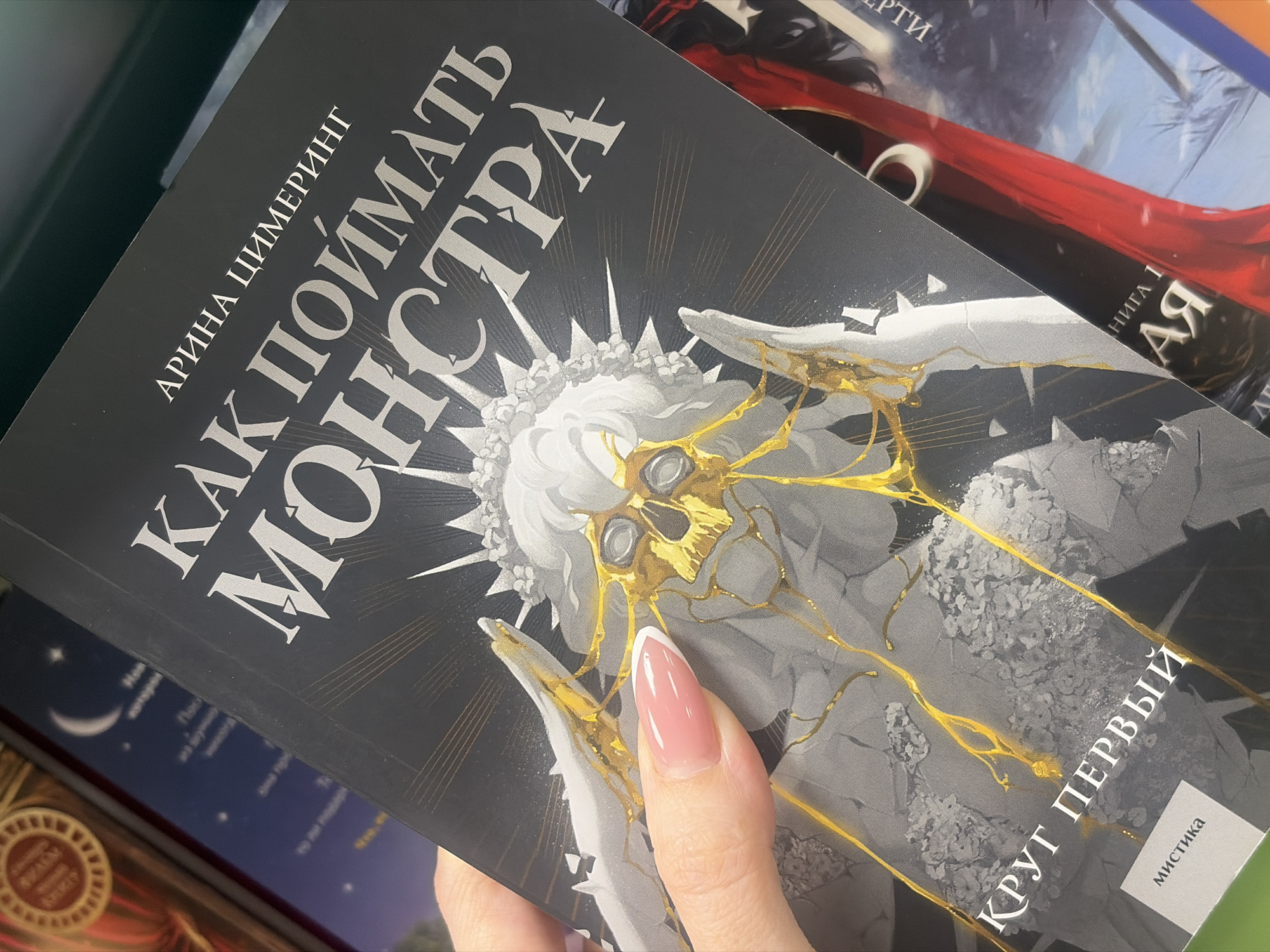 A close-up photograph displays a person's right hand holding a book in an interior setting, identified as a bookstore in Saint Petersburg, Russia. The visible thumb and partial forefinger exhibit a French manicure with pink nail beds and white tips. The held book features a matte black cover with white/silver Cyrillic text. The author, "АРИНА ЦИМЕРИНГ" (ARINA TSIMERING), is printed at the top, followed by the title, "КАК ПОЙМАТЬ МОНСТРА" (HOW TO CATCH A MONSTER). The cover art depicts a stylized, grayscale figure, possibly a robed or skeletal entity, with a spiky crown or halo. Its face is a golden, skull-like mask with empty eye sockets, from which gold liquid or fracture lines extend and drip down the figure's form. Below the main title, "КРУГ ПЕРВЫЙ" (FIRST CIRCLE) and the genre "МИСТИКА" (MYSTICAL FICTION) are visible in smaller text. The background consists of out-of-focus book spines and covers on shelves, including a reddish-brown book with a circular gold emblem, a dark blue book with a crescent moon and stars, and a book with a prominent red spine.
