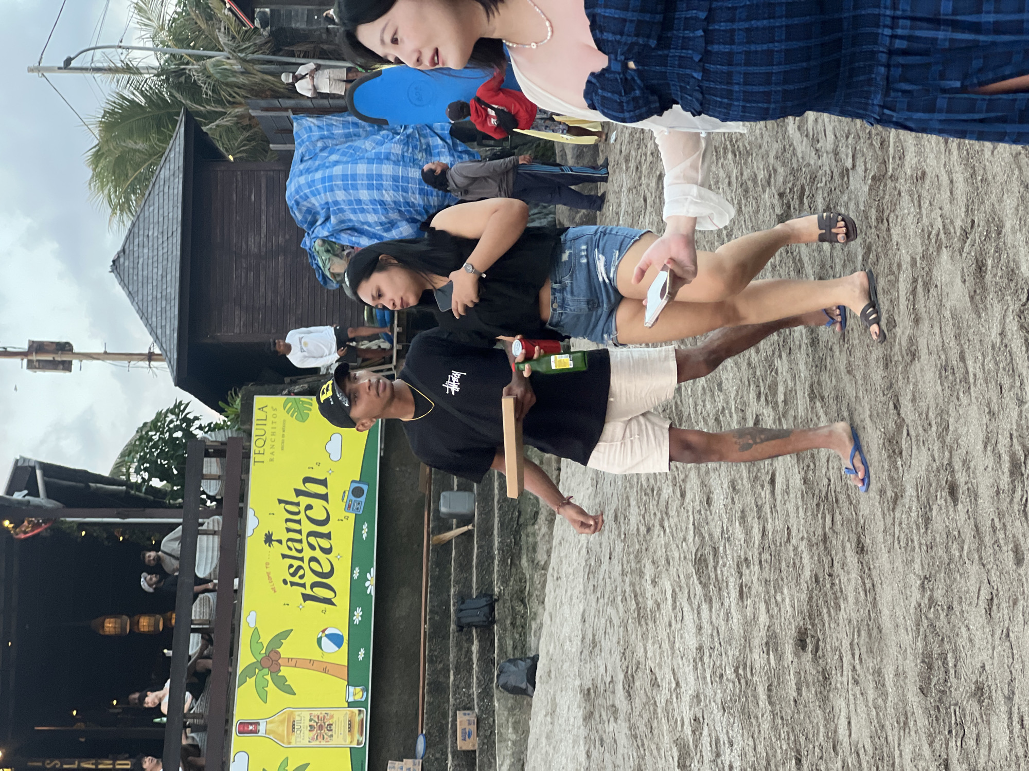 A group of people are on a sandy beach in North Kuta, Indonesia. In the foreground, a man wearing a black t-shirt and beige shorts walks holding a flat, rectangular object and a green bottle. To his right, a woman in denim shorts and a black top walks holding a phone. Further to the right, a woman in a blue plaid dress is partially visible, also holding a phone. In the background, a yellow sign with palm trees and text reads "WELCOME TO ISLAND BEACH". A wooden building with a dark roof and a blue tarp are also visible, along with several surfboards. The sky is overcast.