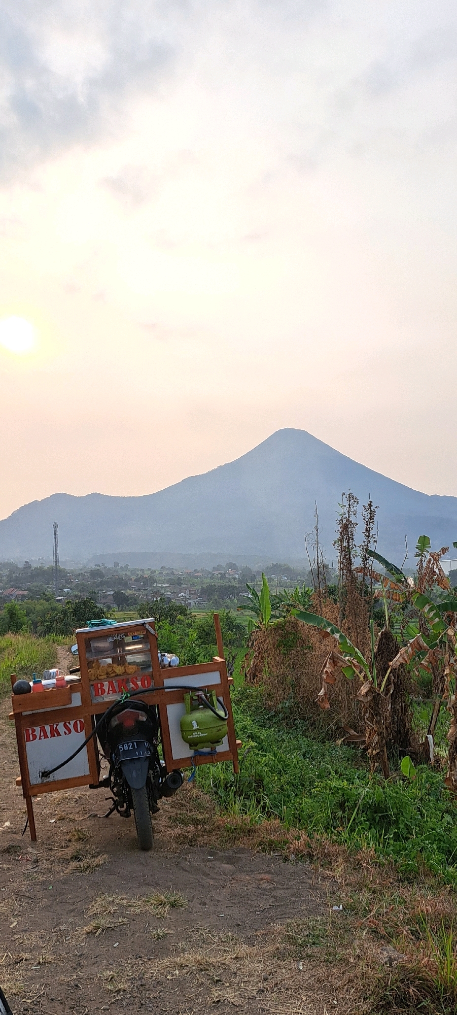 An outdoor, low-angle shot captures a motorcycle-drawn food cart against a backdrop of rolling hills and a hazy mountain range. The sun, partially visible and casting a warm glow, is situated in the upper left quadrant of the frame. The food cart, labeled "BAKSO," is equipped with a green gas cylinder and appears to be set up for business. The surrounding environment is lush with vegetation, including dry and green plants, suggesting a rural or semi-rural setting. In the mid-ground, a village with scattered houses and a prominent telecommunications tower can be observed, with terraced fields visible. The sky is mostly clear with some light clouds. The image was taken in Sumber Gedang, Indonesia.
