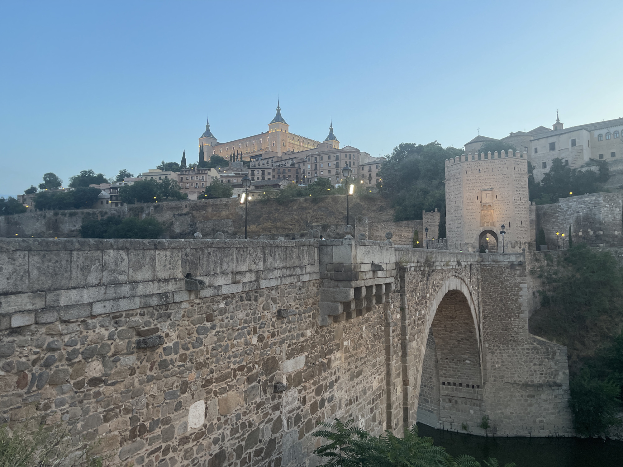 This is an eye-level, medium-shot photograph of the Puente de San Martín in Toledo, Spain, taken at dusk. The stone bridge, a medieval structure, spans a body of dark water, likely the Tagus River, which is visible in the lower right corner of the frame. The bridge's weathered stone construction is detailed, with rough-hewn blocks forming its arches and parapets.
Dominating the background skyline is the Alcázar of Toledo, a large, imposing fortress-palace perched on a hilltop. The Alcázar is illuminated by warm artificial lights, highlighting its towers and crenellated walls. To the right of the Alcázar, a fortified gatehouse, part of the city walls, stands prominently, also lit by yellow lamps. The sky above is a clear, pale blue, transitioning to darker hues at the horizon.
The scene is tranquil, with no visible people. Streetlights are visible along the bridge and the city's elevated structures, casting pools of light. Lush green foliage, likely trees and bushes, frames the lower right edge of the image and creeps up the slopes of the city's hills. The overall impression is one of historic grandeur and a serene evening atmosphere.