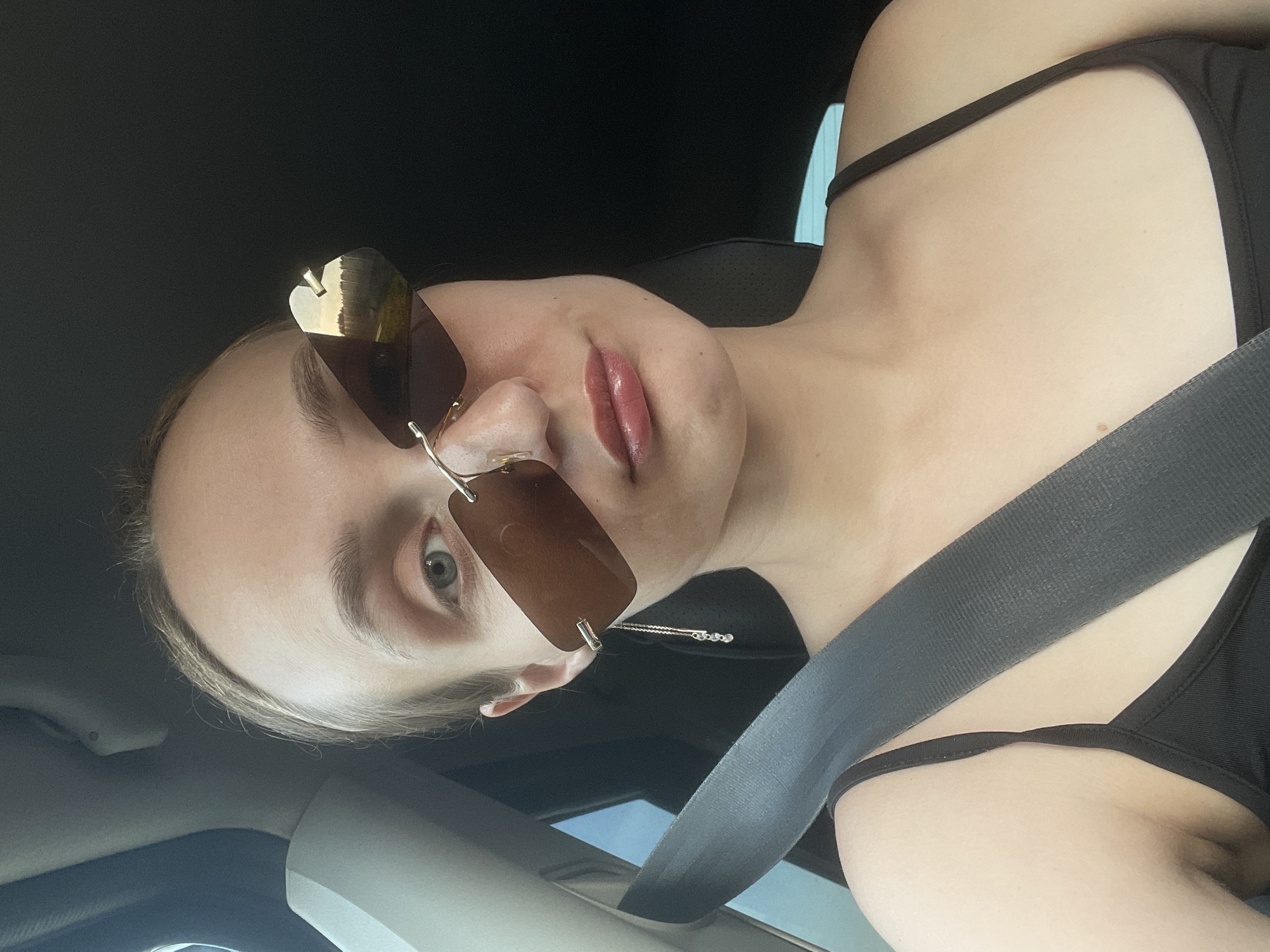 A woman is sitting in a car, wearing sunglasses and a black top, with a seatbelt fastened. The image appears to be taken from a slightly low angle. The location is unknown.