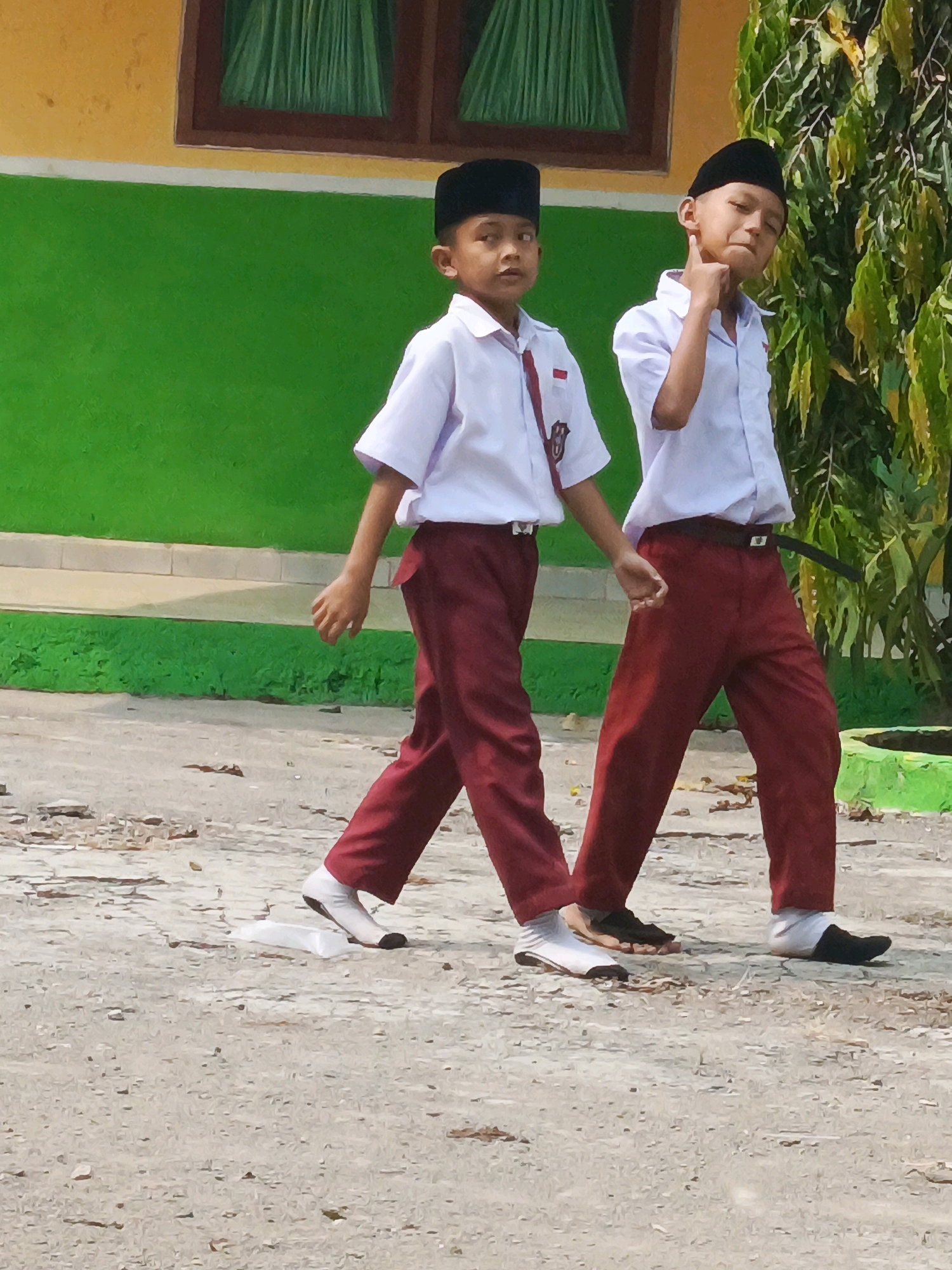 Two young boys, dressed in traditional Indonesian school uniforms consisting of white short-sleeved shirts, maroon pants, black caps, and black shoes with white socks, walk side-by-side on a dirt path. The boy on the left is slightly ahead and to the left of the boy on the right. The boy on the right has his right hand raised, touching his cheek, and appears to be looking to his right. The background features a yellow building with a dark-framed window with green curtains and a vibrant green wall. Lush green foliage is visible to the right of the boys. The ground is a mix of dirt and small stones. The scene suggests an outdoor environment, likely within the school grounds in Karya Dadi, Indonesia.