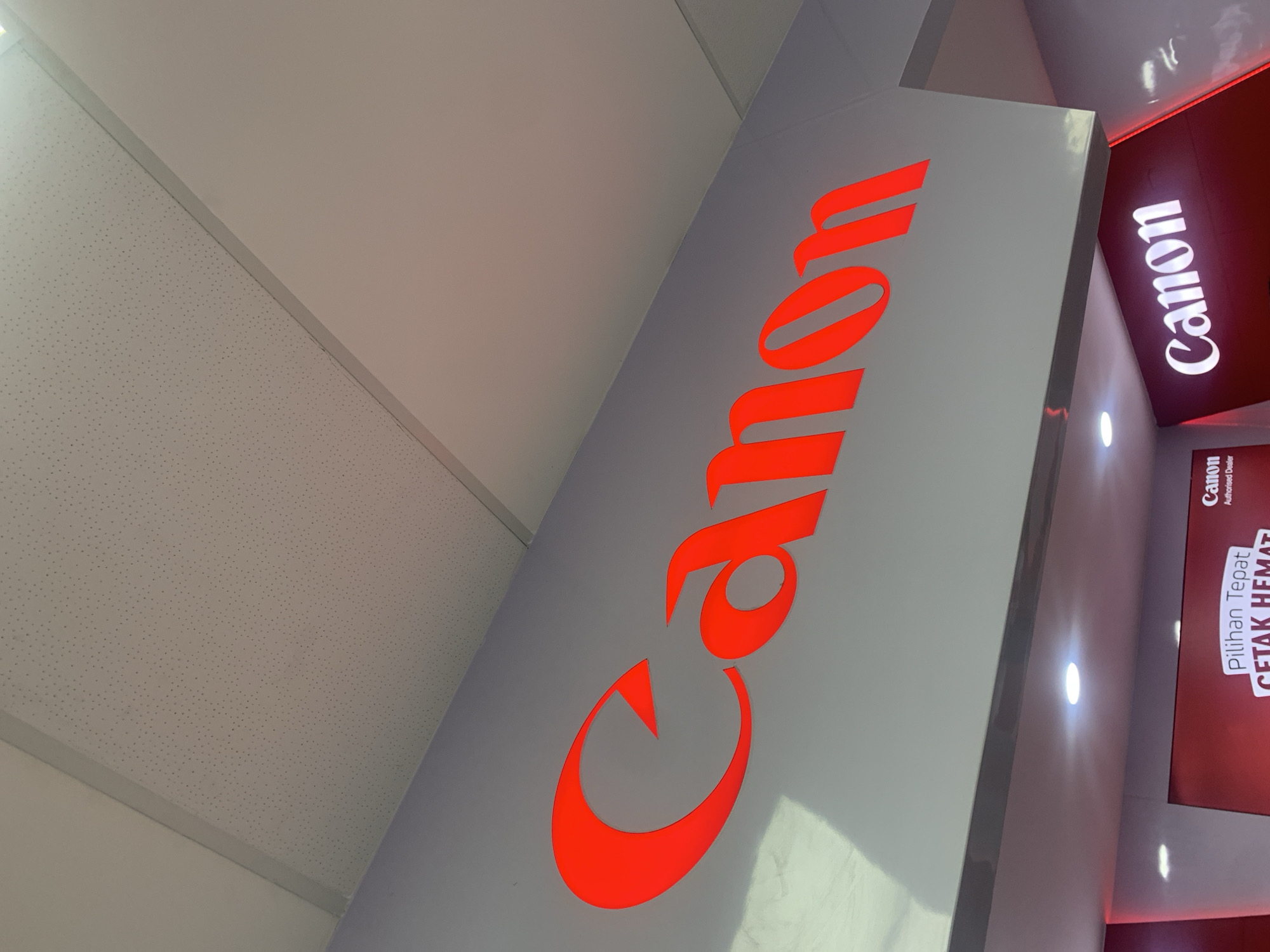 The image shows a brightly lit red "Canon" logo sign mounted on a gray wall, likely in an indoor commercial space. The sign is large and prominent, with the letters appearing to be illuminated from within. Below the main sign, another, smaller "Canon" logo is visible, this one illuminated in white and set against a dark red background, suggesting a display or counter area. Text in a smaller font on a red banner below this second logo is partially visible and appears to be in Indonesian, with the words "Pilihan Tepat" and "CETAK HEMAT" being decipherable, along with "Canon Authorised Dealer". The ceiling above the main sign is a white, textured panel, typical of suspended ceilings in commercial buildings. The lighting suggests the image was taken during the day or in a well-lit interior.
The presence of the "Canon Authorised Dealer" text and the Indonesian language strongly indicate this is a Canon retail store or a designated Canon display area within a larger electronics store in Indonesia. While the specific street address or exact building is not visible, such authorized dealers are commonly found in major shopping malls and commercial centers in cities like Palembang, Indonesia. Therefore, this scene is likely located within a shopping mall or a prominent commercial district in Palembang. There are no people visible in the image.
