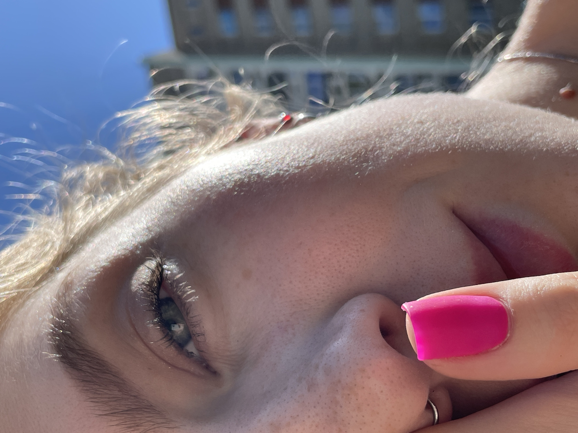 A close-up, low-angle shot focuses on the face of a light-skinned person, with fair hair appearing to be in sunlight. The person's left eye is visible, showing a blue iris. A manicured finger with bright pink nail polish is near the person's nose, and a silver nose ring is visible. The background shows blurred buildings and a blue sky. The location in the city of Izhevsk, Russia is not identifiable from the given image.
