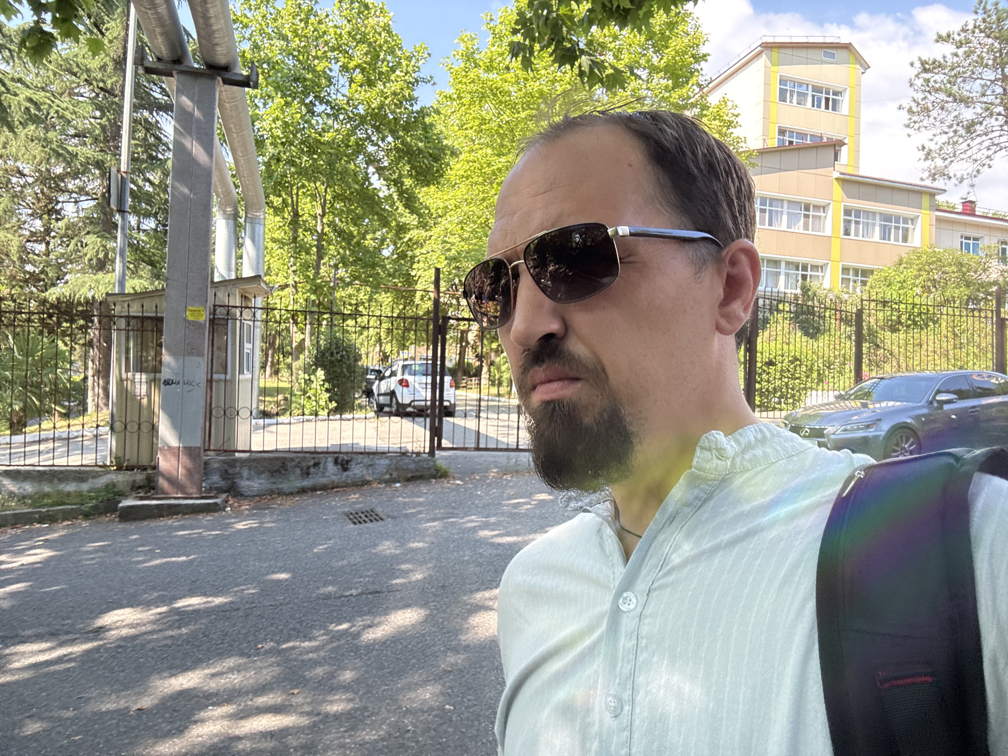 In Sochi, Russia, a man with sunglasses and a beard is standing in front of a school. The school is a multi-story building with a yellow facade and white windows. A metal fence separates the man from the school. Behind the fence, there are trees and a car. The man is wearing a light-colored shirt and a backpack. The sun is shining, casting shadows on the asphalt road. There are large pipes running overhead, supported by a metal structure. A small, booth-like structure is visible near the fence.