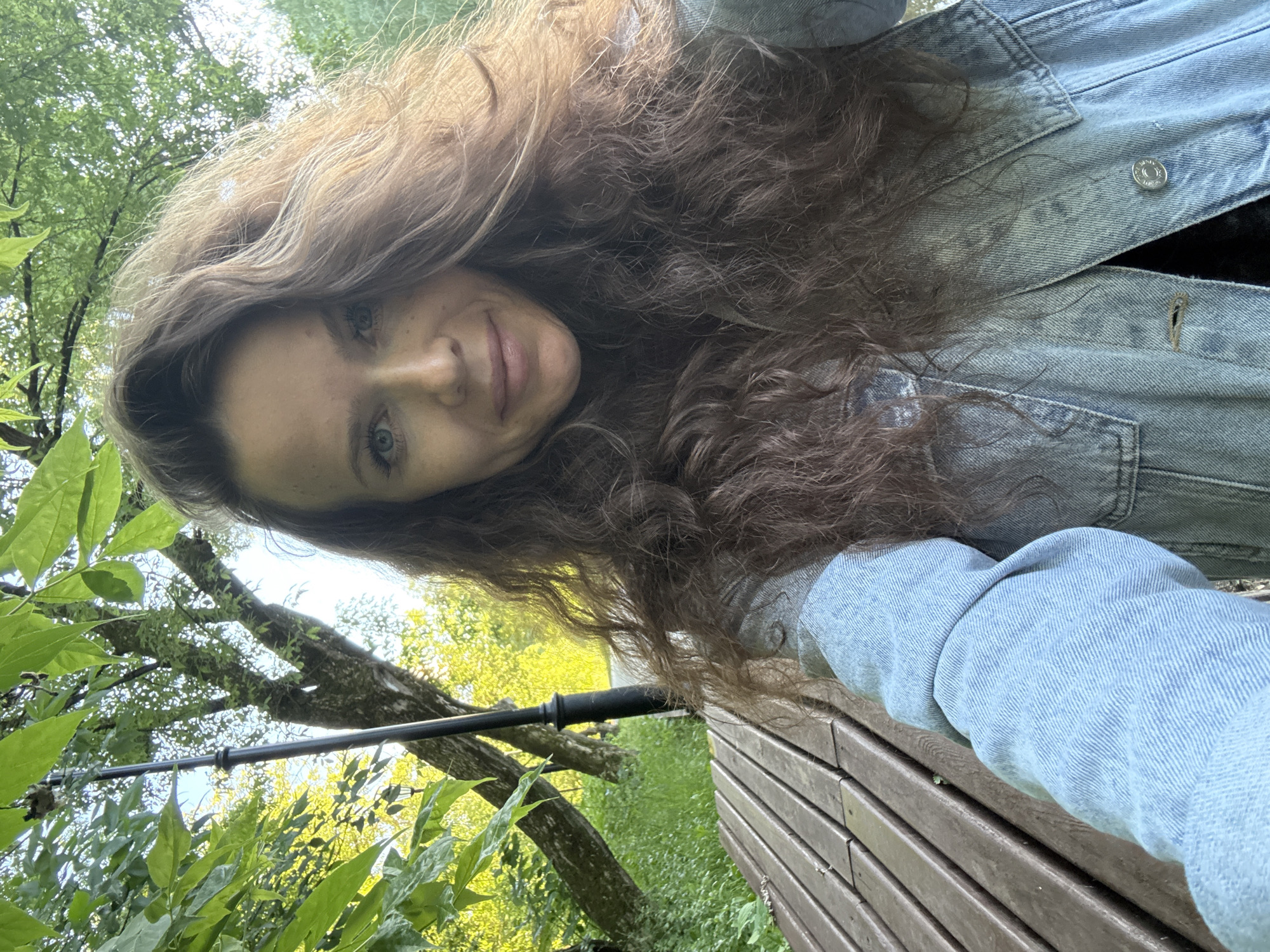 A young woman with long, wavy brown hair is taking a selfie outdoors in a park in Moscow, Russia. She is wearing a light blue denim jacket. Her face is turned towards the camera, and she is smiling slightly. Her eyes are blue, and she has a smattering of freckles across her nose and cheeks.
The background is lush and green, with large trees and foliage. Sunlight filters through the leaves, creating dappled light. A wooden bench is visible in the foreground, suggesting she is sitting or leaning on it. A thin, dark pole, possibly part of a camera setup or a walking stick, runs diagonally through the frame from the lower left.
The scene appears to be set during the daytime, likely late spring or summer, given the abundant greenery and bright sunlight. The overall mood is relaxed and casual. There is no visible text in the image.