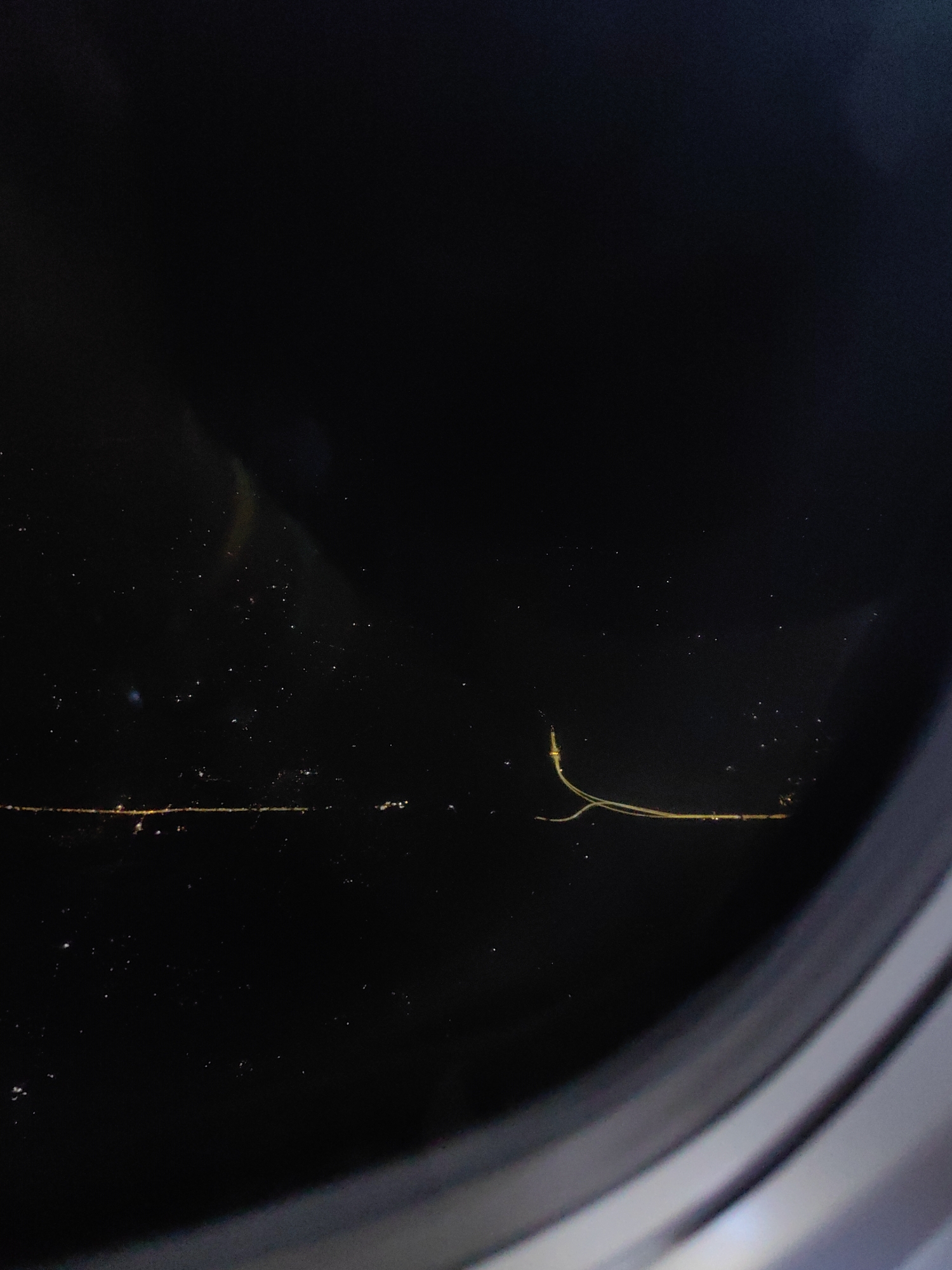 The image shows a dark, nighttime view from an airplane window. The darkness of the night sky is punctuated by scattered lights from the city below, forming a faint, horizontal line. There are also some smaller, more diffuse lights, possibly from distant settlements or stars. A thin, glowing yellow object, possibly a string or fiber, is suspended in the foreground, reflecting the light from the city. The curved frame of the airplane window is visible in the lower right corner, obscuring part of the view. The image does not provide specific information about Paranipur Uparhar, India, or any identifiable people or actions.