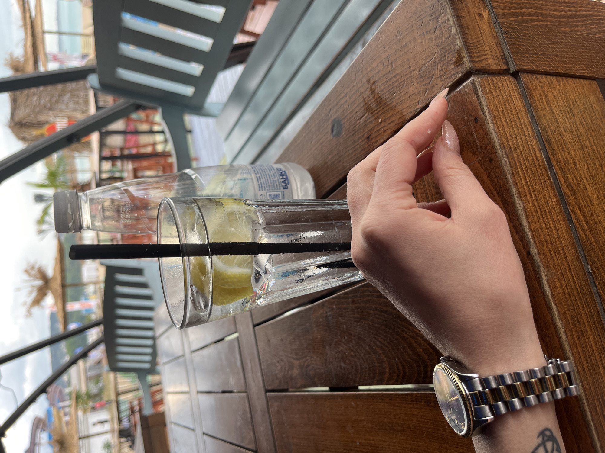 The image depicts an outdoor setting, likely a restaurant or bar, in Varna, Bulgaria. The view suggests a location near the beach, as evidenced by the presence of sand, water, and beach umbrellas visible in the background. A woman's hand and wrist are prominently featured. She is wearing a two-tone metal wristwatch on her left wrist. Her fingers are near the edge of a wooden table, possibly touching it. On the table are a half-empty glass of a drink with lemon slices and a black straw, and a bottle of water. The table is made of wooden planks and has a varnished finish. Part of the seating arrangement includes a chair with a slatted back made of a light-colored material. The orientation suggests the photo was taken at table-level, and the image has been rotated.