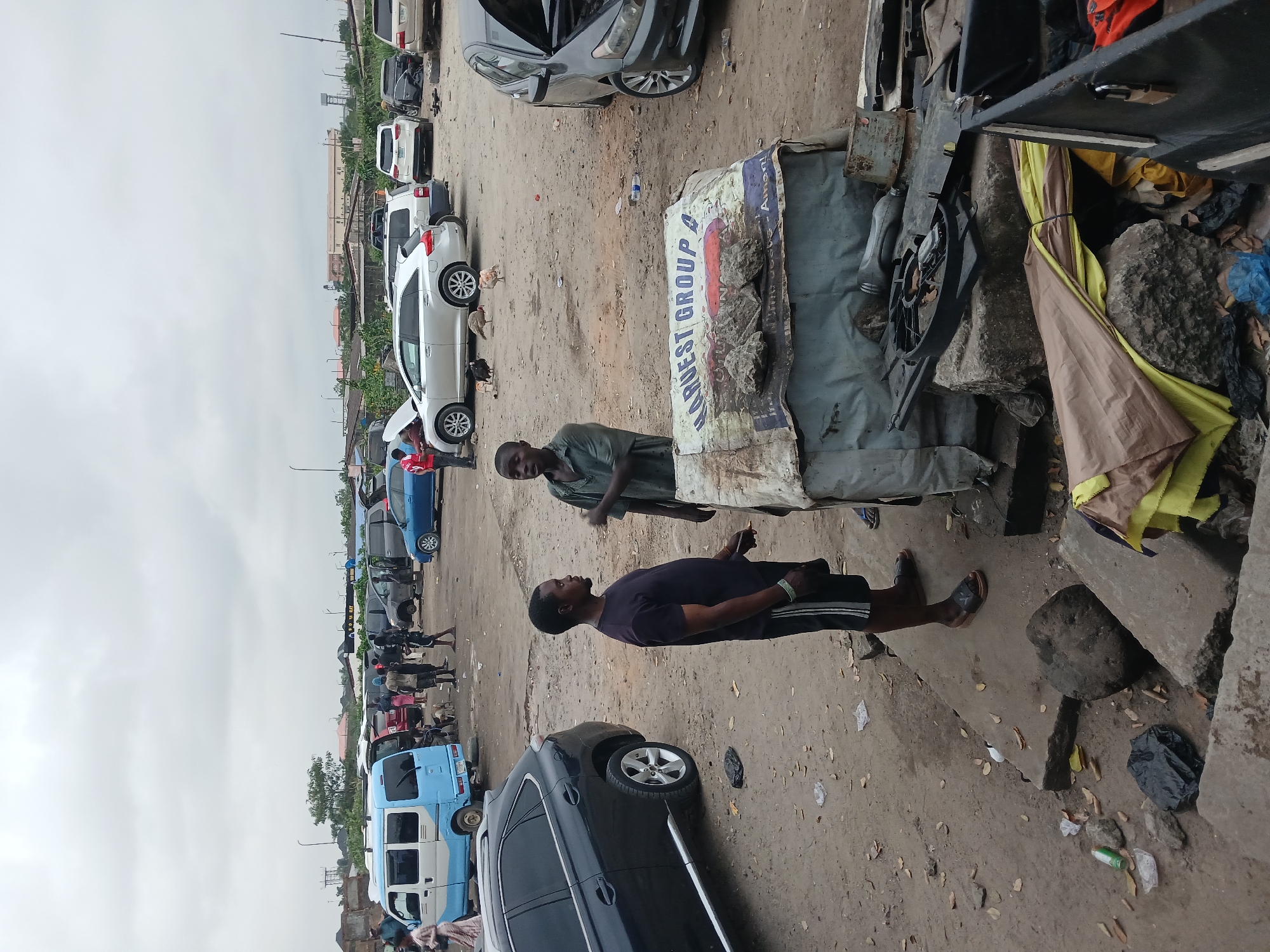 In an outdoor, unpaved area, two men are standing and talking in the foreground. One man, wearing a dark t-shirt and black shorts with white stripes, stands on the left. The other man, wearing a green short-sleeved shirt, stands on the right. They are near a makeshift stall covered with a tarp displaying the text "HARVEST GROUP A". Behind them, a dark SUV is parked. The background shows a collection of parked cars of various types, including a blue and white van, a silver SUV, and a black sedan with its hood open. Several other people are visible in the distance, some standing near vehicles. The ground is covered with dirt, scattered debris, and some fallen leaves. The sky is overcast.