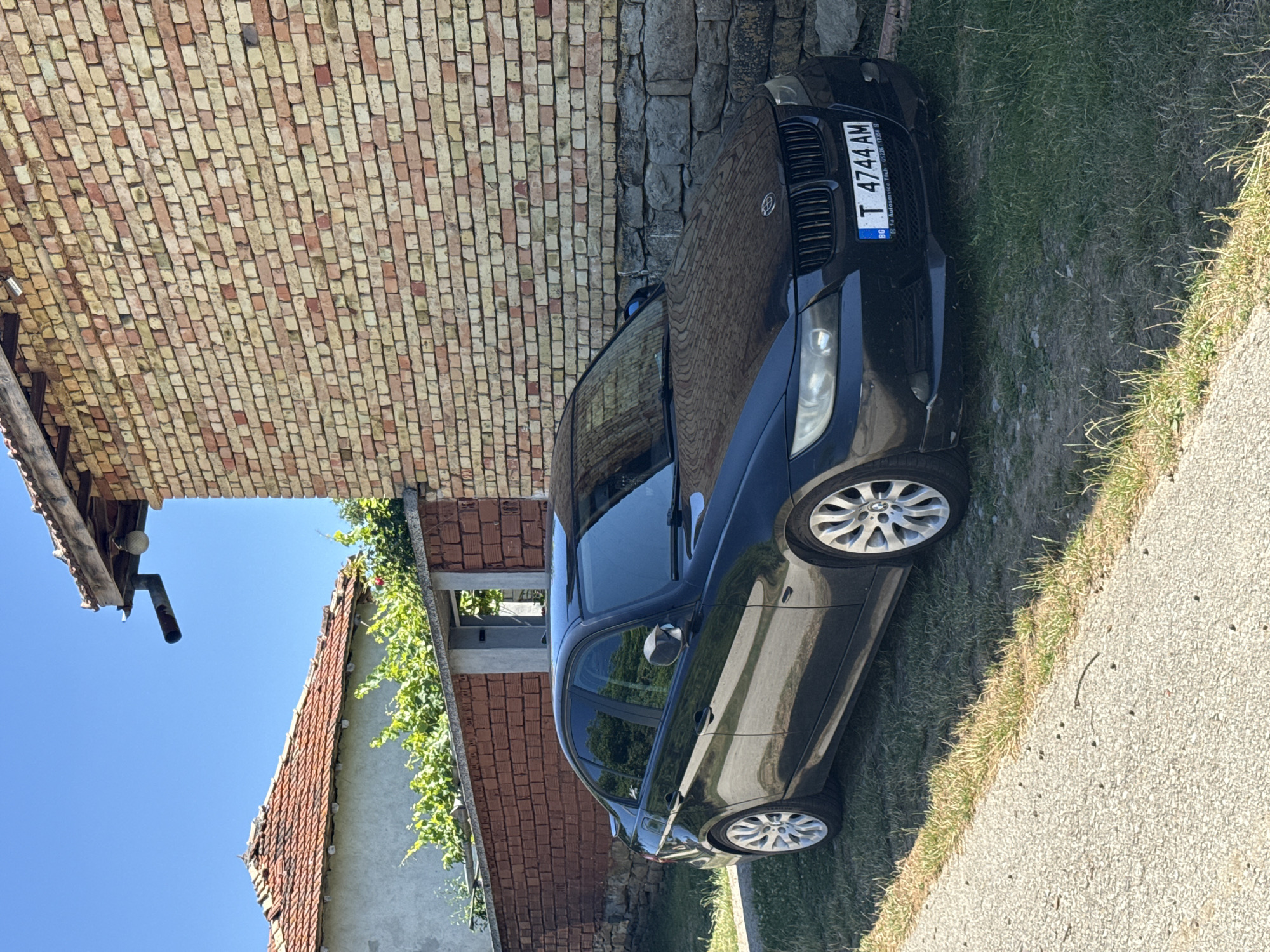 The image shows a black BMW sedan parked on a grassy area next to a gravel path. The car has a Bulgarian license plate that reads "T 4744 AM". Behind the car is a brick wall of a building, and further in the background, there is a tiled roof of another building and some greenery. The sky is clear and blue, indicating a sunny day. There are no people visible in the image. The scene appears to be in a rural or suburban area. Based on the license plate and the general architecture, the location is likely Konop, Bulgaria.