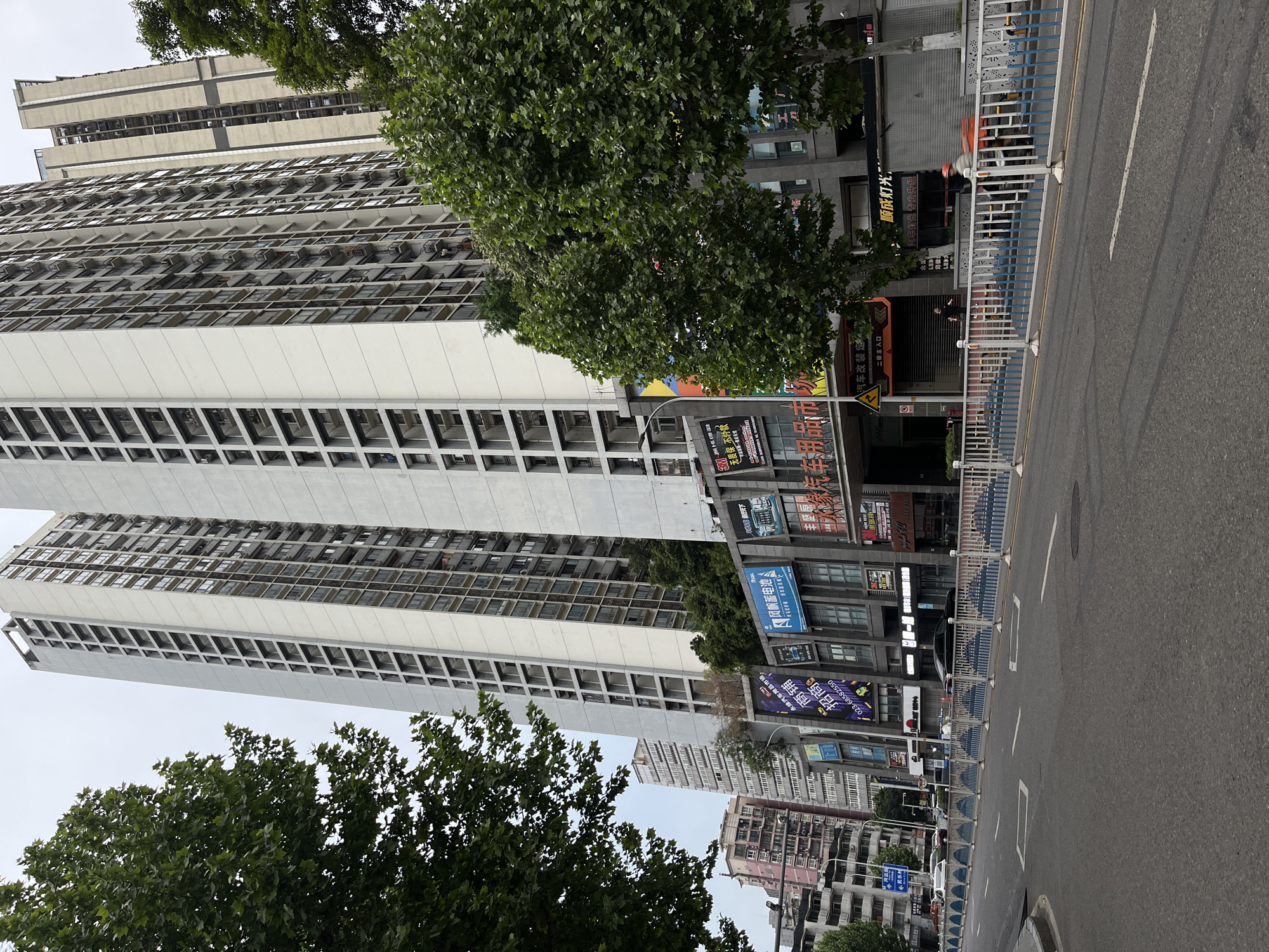 This vertical image captures a daytime urban street scene in Yuzhong District, China, under an overcast sky. Tall, multi-story buildings dominate the view, with many showcasing numerous balconies and external air conditioning units, suggesting residential or mixed-use properties. The lower levels of these buildings feature various commercial storefronts.
Dense green trees are visible in the foreground and midground, partially obscuring the buildings. To the right, a paved road with white lane markings and rectangular ground markings stretches into the distance. A blue and white metal barrier runs along the side of the road, separating it from the buildings and sidewalk area. Behind this barrier, a blurry figure on what appears to be a scooter or bicycle, wearing a helmet and carrying an orange delivery bag, is moving along the street. Two other indistinct figures are visible near a shop entrance further down the road.
Various signs and billboards with Chinese text are prominently displayed on the commercial establishments. Visible text includes: "永缘汽车用品市场" (Yongyuan Auto Supplies Market), "商铺" (Shop), a phone number "023-68582550", "招商" (Investment/Recruitment), "风帆蓄电池" (Sail Battery), "800天下" (800 World), "无质保 不付款" (No warranty, no payment), "汽车改装定" (Car modification customization), "二楼主入口" (Second floor main entrance), "顺成灯光:" (Shuncheng Lighting:), and "汽车灯光专业升级" (Professional upgrade of car lights). The overall impression is one of a busy, functional city street.