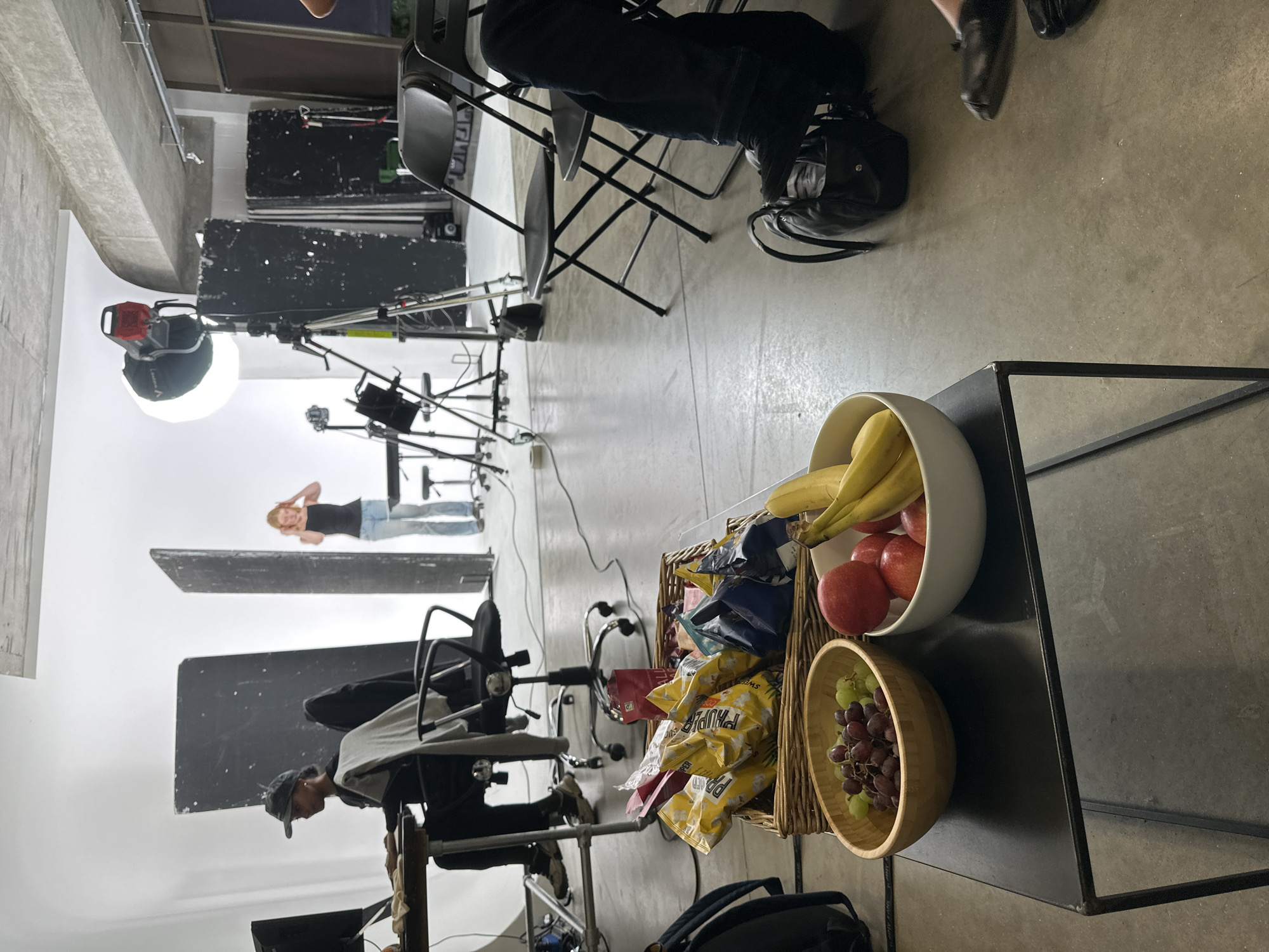 The image shows an interior space, likely a photography or film studio. A fair-skinned female stands in the background, posing in jeans and a black tank top. In the foreground, there's a black metal table holding a white bowl filled with bananas and apples, a wicker basket containing potato chips, and a wooden bowl of grapes. A black folding chair is visible in the midground. Other objects include lighting equipment and dark screens used for setting up the shot. The orientation is rotated. The location is not discernible from the image.