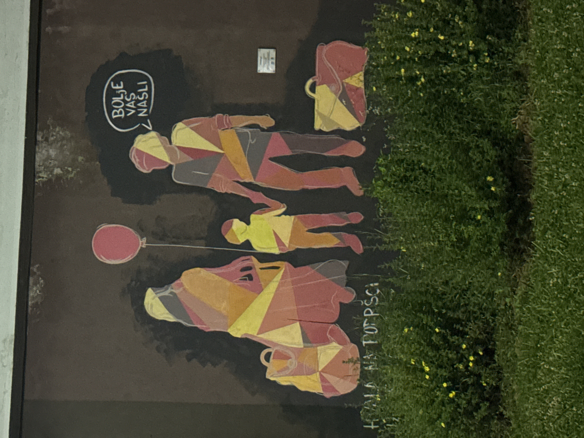 The image shows a mural on a dark brown wall in Ilidža, Bosnia and Herzegovina, depicting a stylized family consisting of two adults and one child, rendered in colorful geometric shapes. The adult figures are holding hands, and the child stands between them. To the left of the family is a large bag, and to the right, another bag. A pink balloon floats above the child. To the upper right of the mural, inside a white speech bubble, are the words "BOLJE VAS NASLI" in white lettering. Below the mural, a sentence in white capital letters reads "HVALA NA POTPISCI". The mural is positioned above a dense green bush with small yellow flowers, and in the foreground, there is a grassy area.