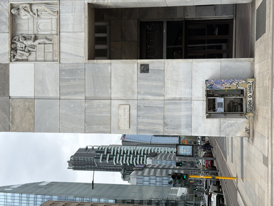 The image displays a street scene on Via Vittor Pisani in Milan, Italy, under an overcast sky. On the right, a building facade constructed from light grey, rectangular stone panels features a stone relief sculpture in the upper right, depicting multiple figures including three males, one actively pulling a rope, and a standing female figure. A rectangular plaque on the facade, centered left, identifies "VIA VITTOR PISANI". Below this, a recessed entry with dark brown double doors and an overhead window is present. At the base of the facade, an open utility box exposes internal components; its exterior is marked with purple bubble graffiti and the phrase "SUGO O VINO?" written in black at the bottom.
To the left, a modern urban environment is visible. Multiple high-rise buildings, characterized by glass and steel construction, including one with prominent white, tiered balconies, rise into the background. A yellow pole supports a green traffic light for vehicles and a distinct green bicycle signal. A paved sidewalk extends along a street occupied by numerous parked and moving cars. Several indistinct pedestrians are observable further down the street. A streetlamp and an advertisement display are also present within the scene.
