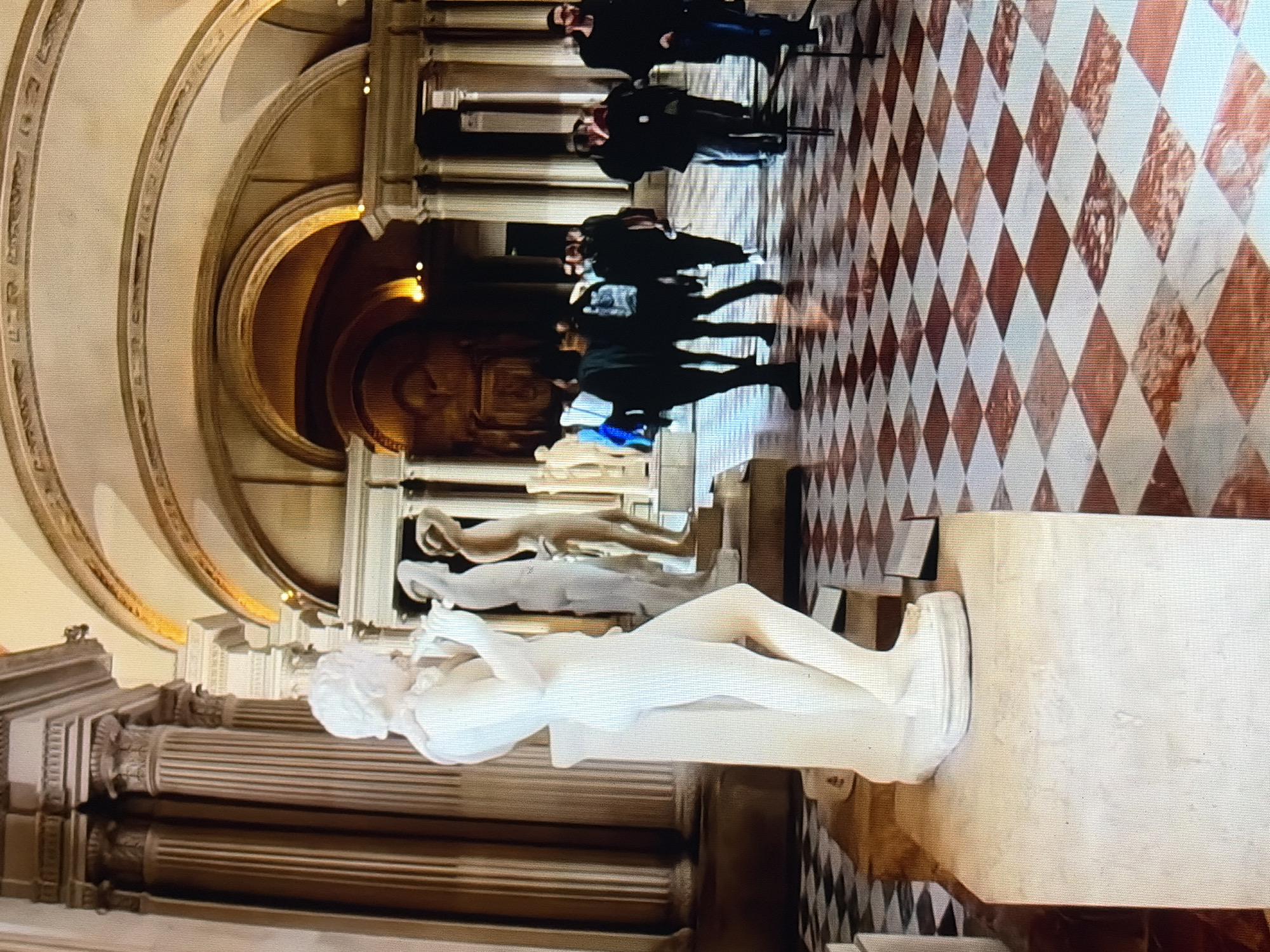 The image shows the interior of a museum, possibly an art gallery, with classical architecture. In the foreground, a white marble statue of a youth playing a flute stands on a pedestal. The statue is a classical sculpture, rendered in white marble with a smooth finish. Behind the statue, other classical sculptures are visible, including what appears to be a draped figure and another male form. The floor is a checkered pattern of white and reddish-brown marble tiles, arranged in diamonds. In the background, several people, dressed in casual clothing, are walking through the museum. The architecture features grand columns, arched ceilings, and ornate detailing, suggesting a historical building. The lighting is ambient, with some spotlights highlighting the artworks. The overall scene conveys a sense of quiet observation and cultural appreciation.