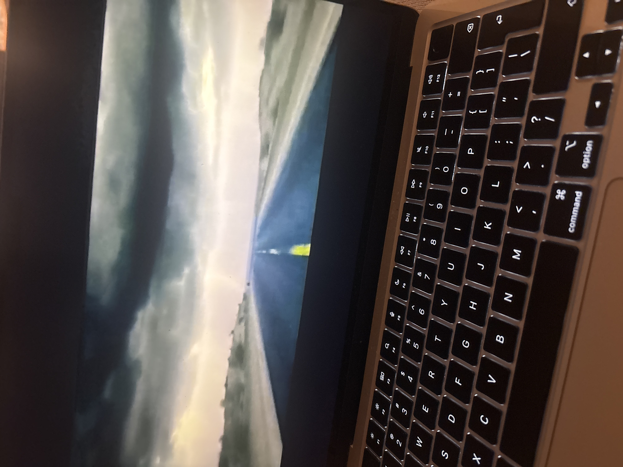 A close-up, angled shot captures a laptop screen displaying a landscape image of a road stretching towards a stormy sky. The road is paved with a yellow line down the center, bordered by a dry, barren landscape. Dark, dramatic clouds fill the upper portion of the screen. The laptop's illuminated keyboard is visible in the foreground, with keys such as "Q", "W", "E", "R", "T", "Y", "U", "I", "O", "P", "A", "S", "D", "F", "G", "H", "J", "K", "L", "Z", "X", "C", "V", "B", "N", and "M" clearly legible. Function keys F1 through F12 are also visible. The laptop appears to be on a wooden surface. The image does not contain any information about the city of Sofia, Bulgaria.