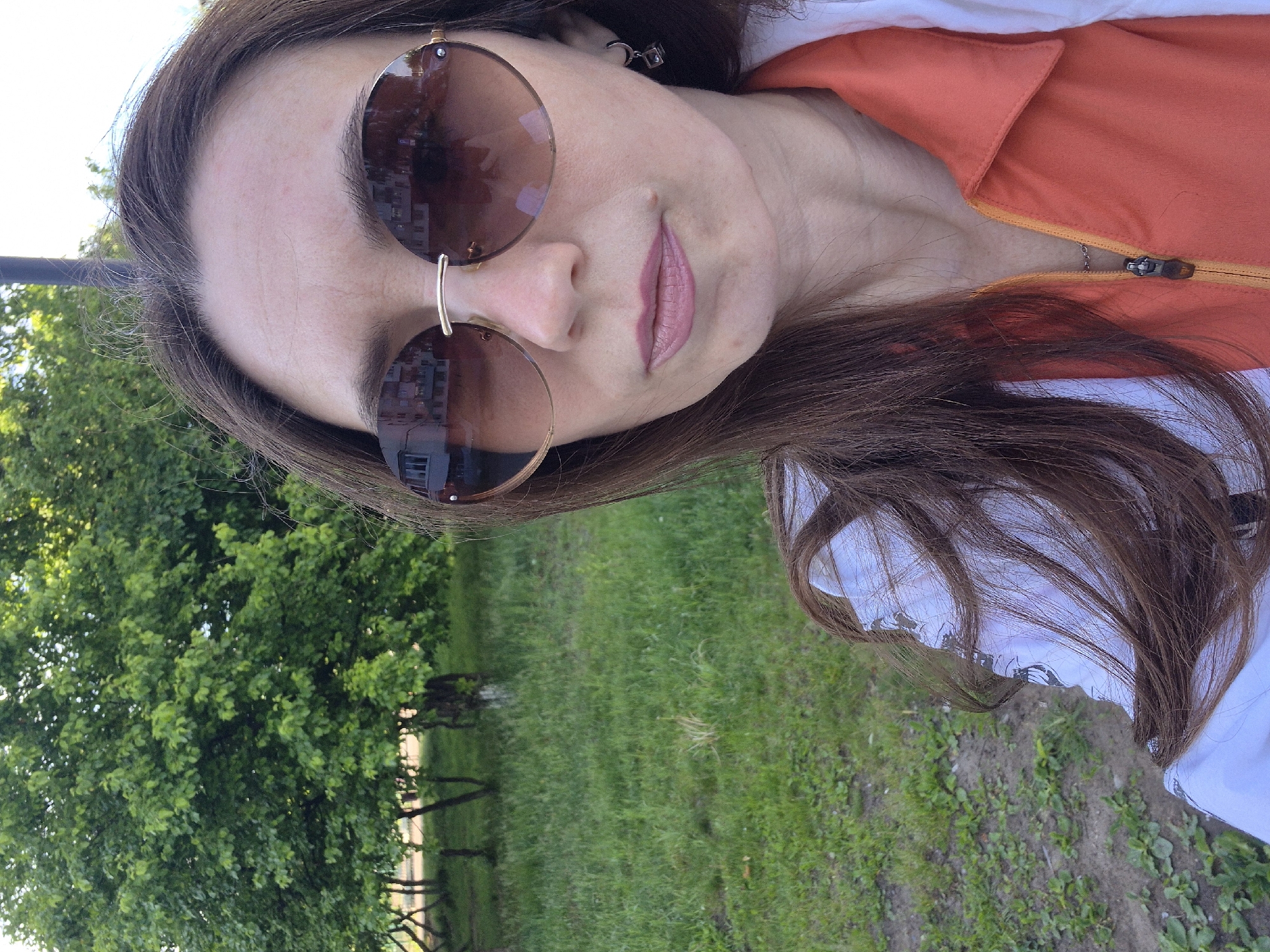 This image is a portrait-style shot of a person, likely female, taken outdoors during the daytime. The photo is rotated 90 degrees clockwise, showing the subject's head on the left side of the frame.
The subject has long, dark brown hair and is wearing large, round sunglasses with brown-tinted lenses and a slender gold-colored frame. Reflections in the sunglasses reveal glimpses of buildings and trees, indicating an urban or suburban outdoor environment. Her lips are painted with a light pink or nude lipstick, and a subtle smirk is visible. A small mole is noticeable above her upper lip. A square, metallic earring is visible on her right ear (viewer's left).
She is wearing an orange collared top, possibly a jacket or shirt, which has a silver zipper with yellow tape. Underneath the orange top, a white garment is visible. The background features a vibrant green landscape, dominated by lush trees with dense foliage and a grassy ground. There are also patches of bare soil visible. The bright, clear lighting suggests it is a sunny day. No visible text is present in the image.