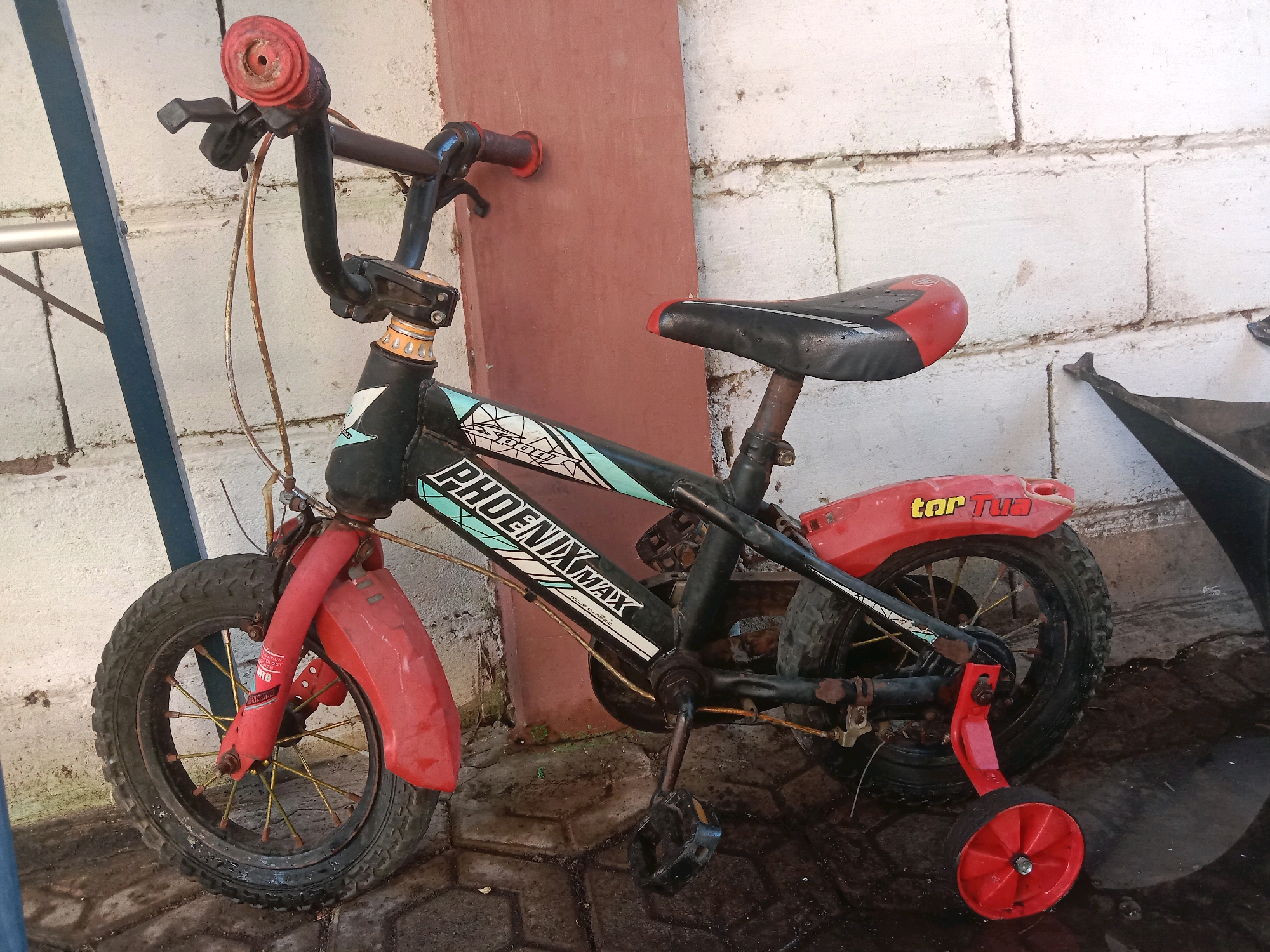 An outdoor, eye-level medium shot captures a child's black and red Phoenix Max bicycle with training wheels parked on a wet, tiled ground against a white brick wall and a reddish-brown wall. The bicycle is positioned at a slight angle, with the front wheel pointing left and the back wheel pointing right. The bicycle's handlebars are black with red grips, and a brake lever is visible on the left. The seat is black with red accents. The frame of the bicycle is black with green and white decals that read "PHOENIX MAX". Red fenders are present on both the front and back wheels. The front wheel has thin, yellow spokes. The training wheels are red. The ground appears to be paved with square tiles and is wet, with some dark areas suggesting puddles or dampness. The background consists of a white brick wall with visible mortar lines and a portion of a reddish-brown wall. To the left of the bicycle, a blue metal support pole is partially visible. The lighting suggests it is daytime, with soft shadows.