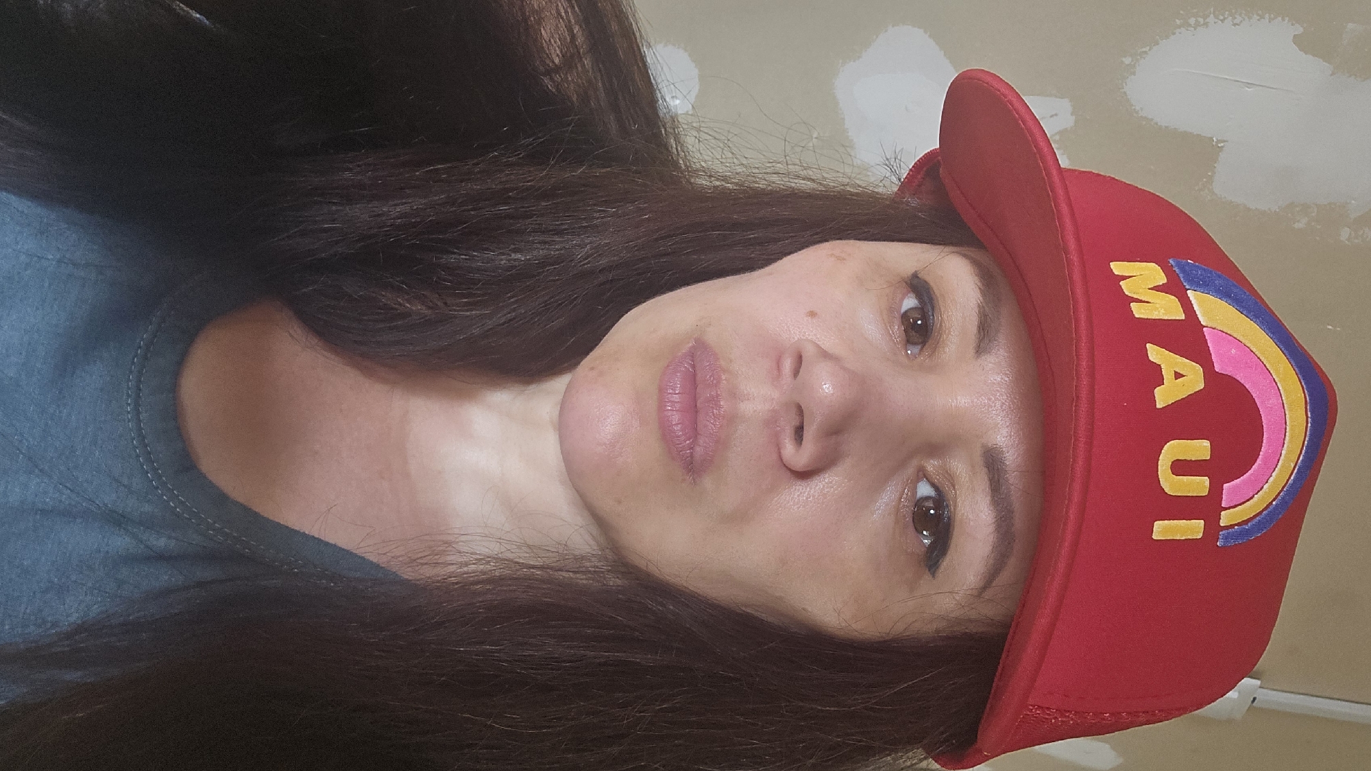A medium close-up selfie shows a woman wearing a red cap with a rainbow and the word "MAUI" embroidered on it. She has long, dark hair and is looking directly at the camera with a neutral expression. Her makeup includes eyeliner and lip gloss. She is wearing a blue t-shirt. The background appears to be a wall with patches of white plaster. There is no indication of the image being taken in Ермолаево, Russia.