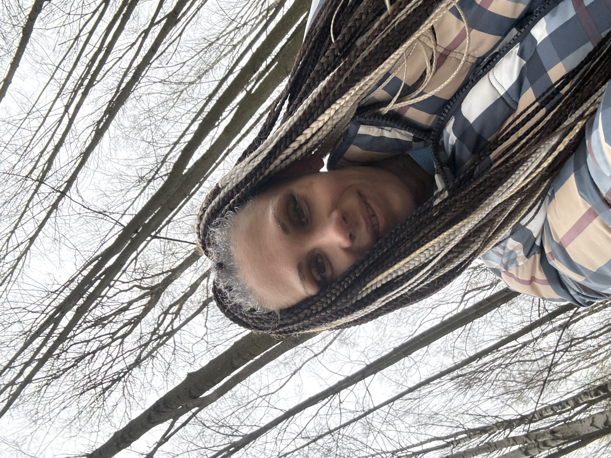 An upward-angled outdoor portrait captures a woman's face and upper torso, framed by bare tree branches against an overcast sky. The woman, positioned in the lower part of the frame, looks directly at the camera with a soft, gentle smile. Her hair is styled in long, intricate braids or dreadlocks, featuring a multi-toned blend of dark brown, black, and prominent light gray or white strands that frame her face and extend downwards.
She is wearing a quilted jacket with a distinct plaid pattern in shades of blue, white, and a muted beige or light brown, suggesting cool weather attire. A glimpse of a solid blue garment is visible beneath her jacket.
The background consists of numerous leafless tree branches and trunks, reaching upwards and crisscrossing against a bright, diffused white sky, indicative of an overcast day during late autumn, winter, or early spring. The scene suggests a wooded area, park, or forest, consistent with the natural landscape of Rusanovo, Russia. The low angle implies the photo was taken from below, possibly a selfie, with the woman looking up. No visible text is present.