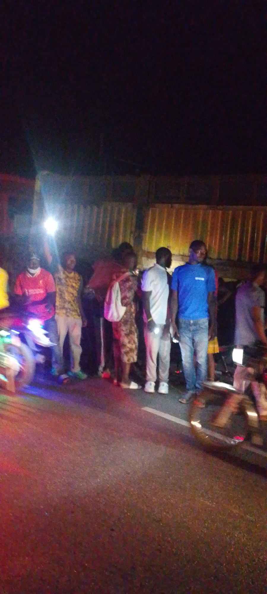 A group of approximately eight people stands on a paved road at night, observed from a low angle. The location appears to be in a city, possibly Unknown, Unknown, given the street surface and nearby corrugated metal structure. One individual holds a light source aloft, illuminating the scene. The people are casually dressed in a mix of t-shirts, casual pants, and footwear including sneakers and sandals. The road shows signs of wear and a white line marking. A motorcycle is present on the left of the frame, emitting a blue and purple light. Another motorcycle appears on the right, partially visible. The background is dark with limited visibility. The people are mostly standing, seemingly observing or discussing something outside the frame.