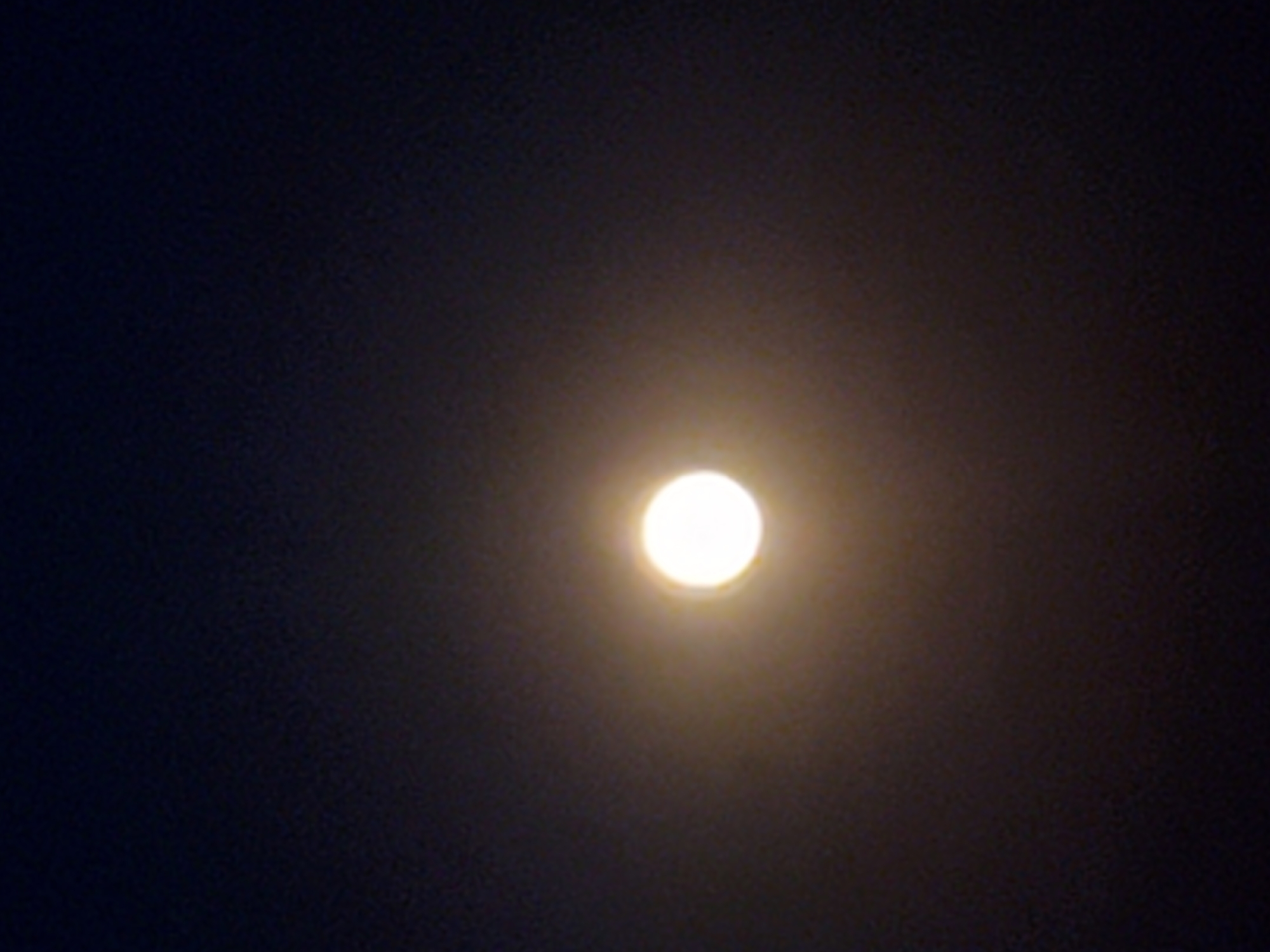The image is a vertical, low-light photograph of a bright, luminous full moon against a dark night sky. The moon is positioned near the center of the frame and is the primary subject. It is a bright, yellowish-white orb with a soft halo effect due to overexposure or atmospheric conditions. The sky is a deep, muted blue-black, with subtle variations in shade suggesting depth. There are no discernible details of the landscape or any urban features of Konstantinovka, Moldova, visible in the image. The focus is solely on the celestial body.