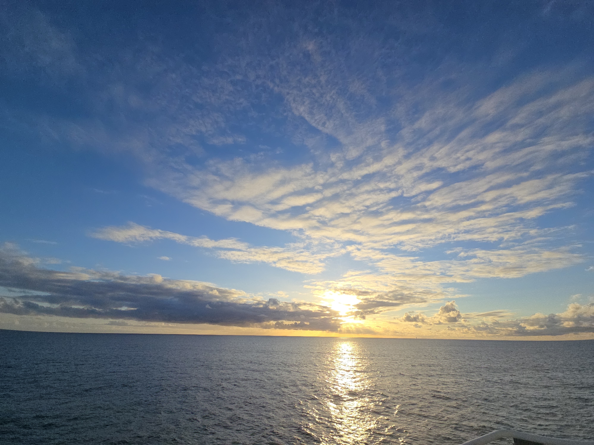 The image depicts a seascape at sunset. The sky is predominantly blue with streaks of white and gray clouds scattered across it. The sun is positioned near the horizon, partially obscured by the clouds, creating a bright golden glow and a reflection on the water's surface. The sea is calm with gentle ripples and a gradient of color from dark blue to golden where the sunlight reflects. There are no people or identifiable objects in the image besides the natural elements. The location is unknown.