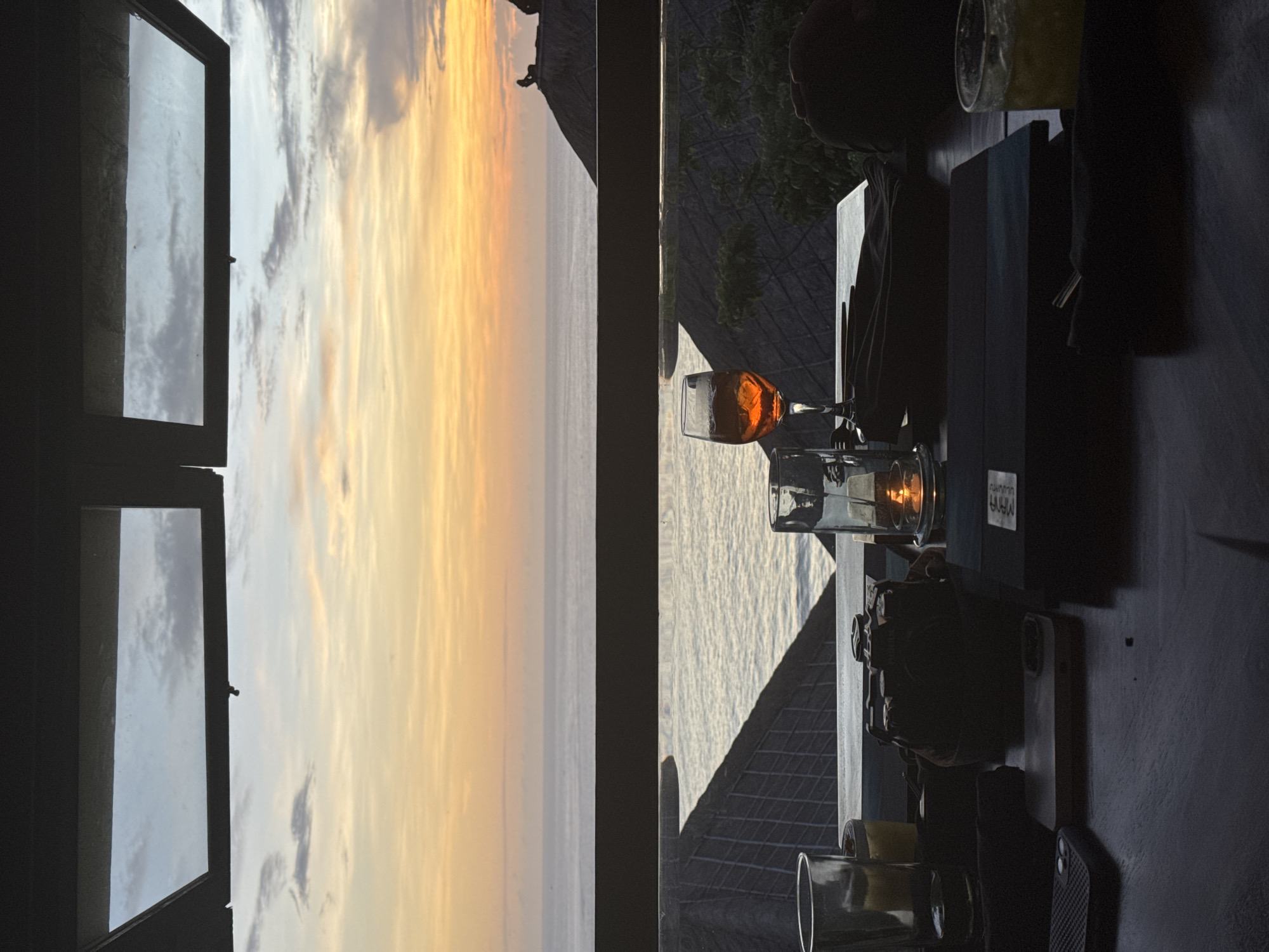 The image depicts a scene from what appears to be a restaurant or bar, with a view of a sunset over the ocean through a window. The interior is dimly lit, and there are various objects on the table including glassware containing liquids (possibly alcoholic beverages), serving trays, and personal items such as phones. The window is partially open, revealing the sky painted with hues of orange, yellow, and gray clouds. The scene is located in an unknown city in an unknown location.