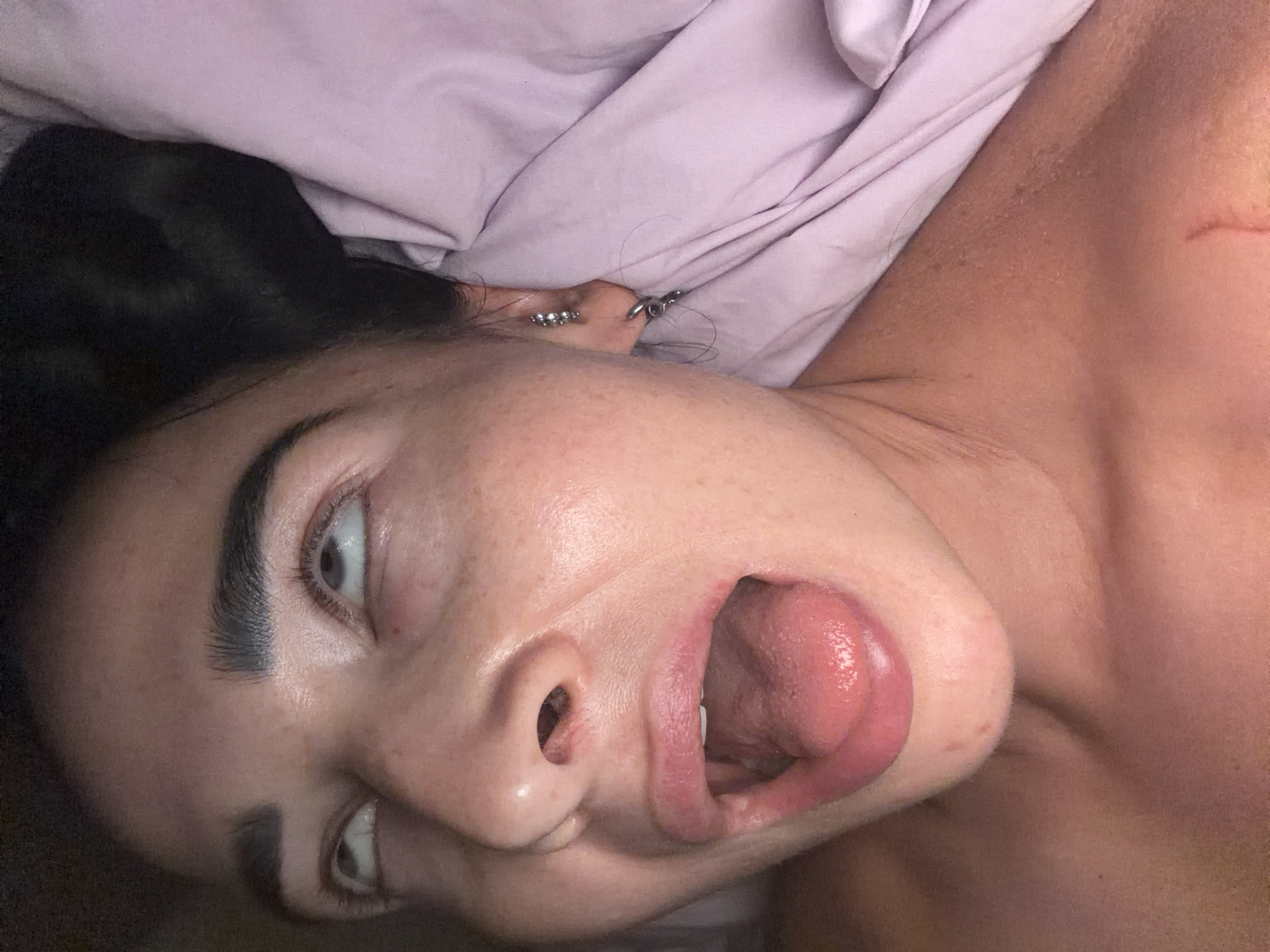 A close-up, rotated portrait captures a person, likely female, lying down, partially covered by a light purple blanket or sheet. The individual has dark hair and a fair skin tone. Their face is the central focus, featuring wide-open eyes looking upwards and an open mouth with the tongue slightly extended. The expression conveys a sense of playfulness, silliness, or surprise.
The visible ear is adorned with multiple silver-toned piercings, including studs and a hoop. The person's eyebrows are dark and well-defined. On the exposed shoulder and neck area, a reddish mark or scratch is noticeable on the skin, which also exhibits natural texture. The setting appears to be indoors, possibly a bedroom, under soft lighting. There is no visible text in the image. This scene is located in Saint Petersburg, Russia.