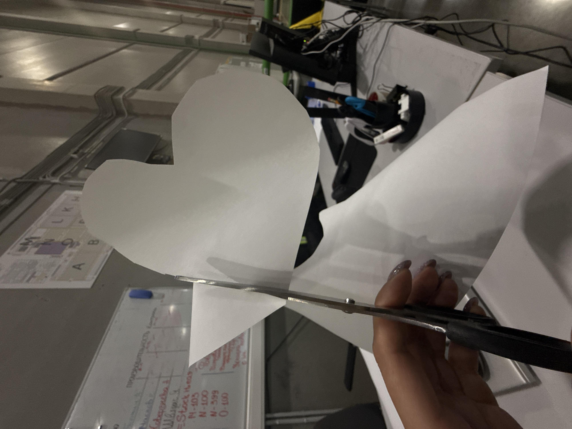 A person is using scissors to cut a white heart shape out of a sheet of white paper. The scene is indoors, likely an office or workshop environment, as indicated by the whiteboards with text in what appears to be Russian, and other office supplies visible on the white desk. There are no landmarks or signs indicating that the location is Lupanovo, Russia. The ceiling is unfinished.