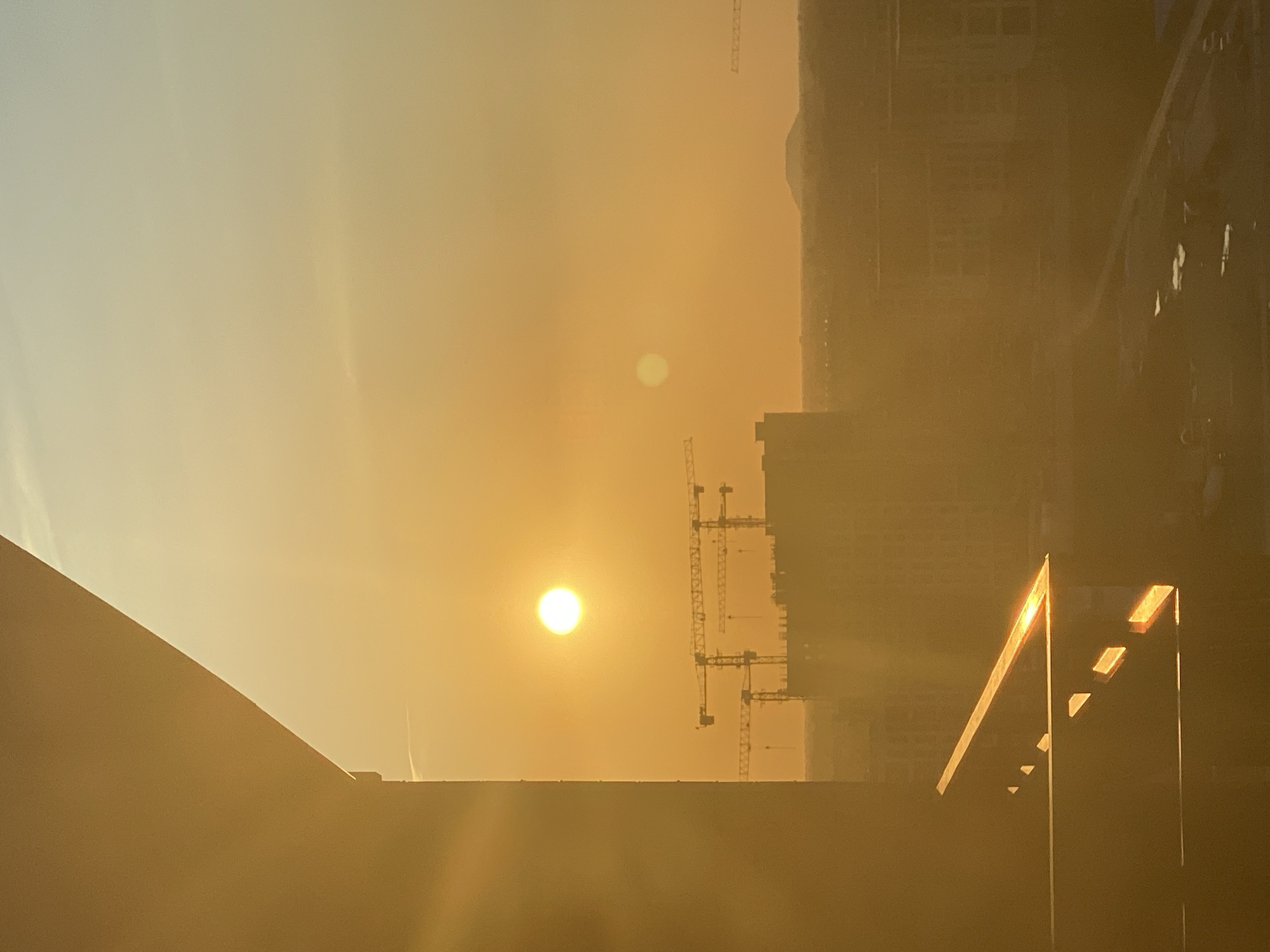 The image captures a sunset over an urban landscape. The sun, a bright orb, is positioned in the upper-middle section of the frame, casting a warm, golden hue across the sky and the scene below. In the foreground, a portion of a building's structure is visible on the left, its angled surface illuminated by the setting sun. A balcony railing with distinct reflections of the sunlight is also present.
In the mid-ground, several construction cranes are silhouetted against the bright sky, indicating ongoing development. Behind the cranes, a cluster of buildings forms the city's skyline, appearing somewhat indistinct due to the atmospheric haze and the intensity of the sunset. Further in the distance, a faint outline of a hill or mountain is visible on the horizon. The sky transitions from a bright golden orange near the sun to a lighter yellow and then a pale blue towards the top of the image. The overall impression is one of a peaceful, yet active, urban environment at dusk.