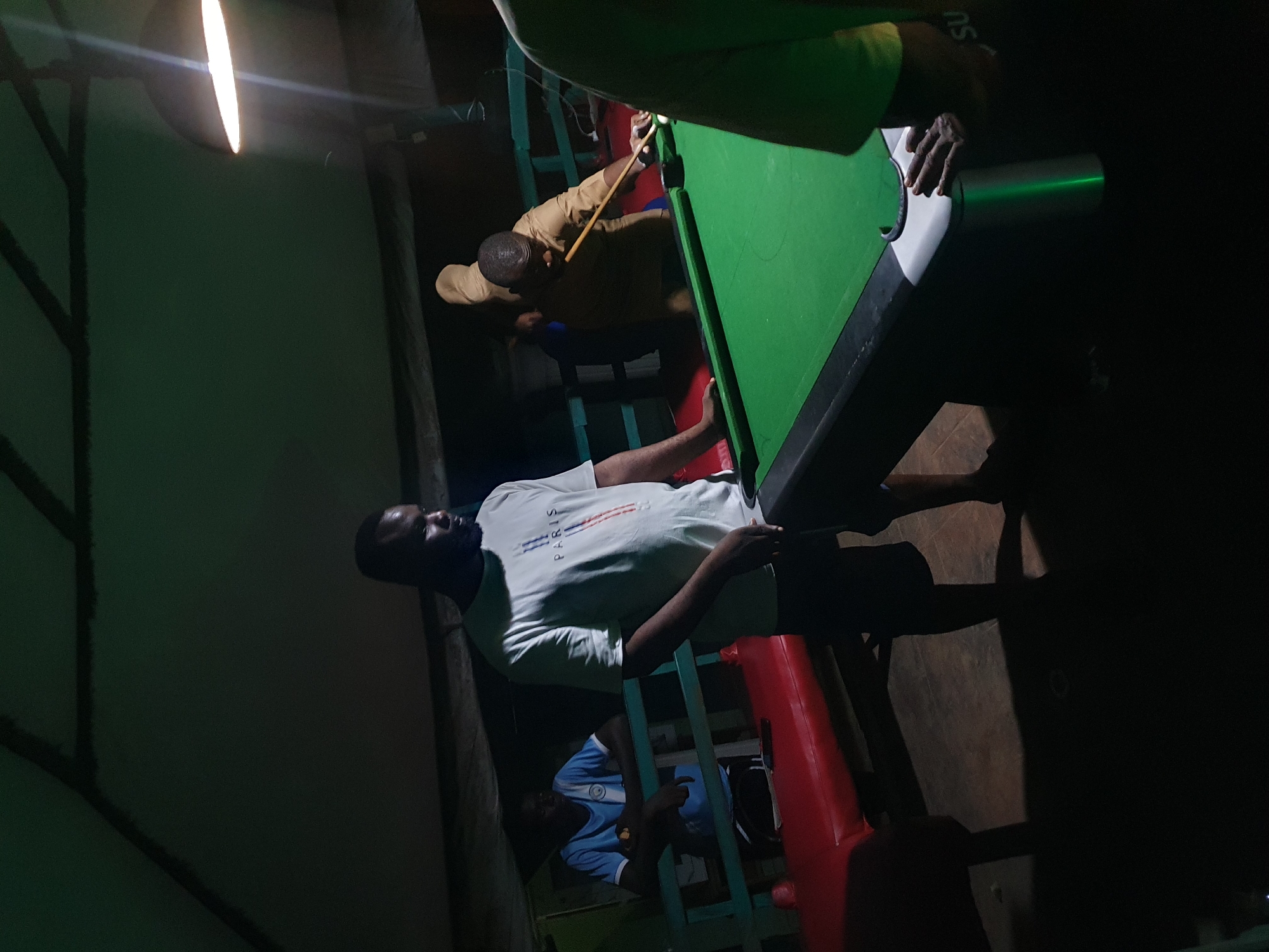In Lokoja, Nigeria, a group of men are engaged in a game of pool in a dimly lit, indoor establishment. The scene appears to be a recreational area, possibly a bar or lounge, with a pool table as the central focus. The table is covered in green felt, and the surrounding area is dark, with a single overhead light illuminating the game.
One man, wearing a white t-shirt with "PARIS" printed on it, stands close to the pool table, looking down at the game. He has a beard and appears to be observing the play. To his right, another man, dressed in a light brown shirt, is actively playing pool. He is in the process of taking a shot, holding a pool cue and aiming at a white ball on the table. A third man, wearing a light blue t-shirt, is visible in the background, seated on a bench behind the pool table, also observing the game. The bench is red and appears to be made of some sort of cushioned material.
The environment is rustic, with exposed beams and a ceiling that suggests a simple construction. The floor is dark and appears to be made of concrete or a similar material. The overall impression is one of casual social gathering, with the pool game providing a focal point for the men's interaction. The presence of a light fixture hanging from the ceiling, which is partially visible, further indicates an indoor setting. The shadows cast by the people and the pool table suggest the intensity of the light source and the low ambient light conditions.