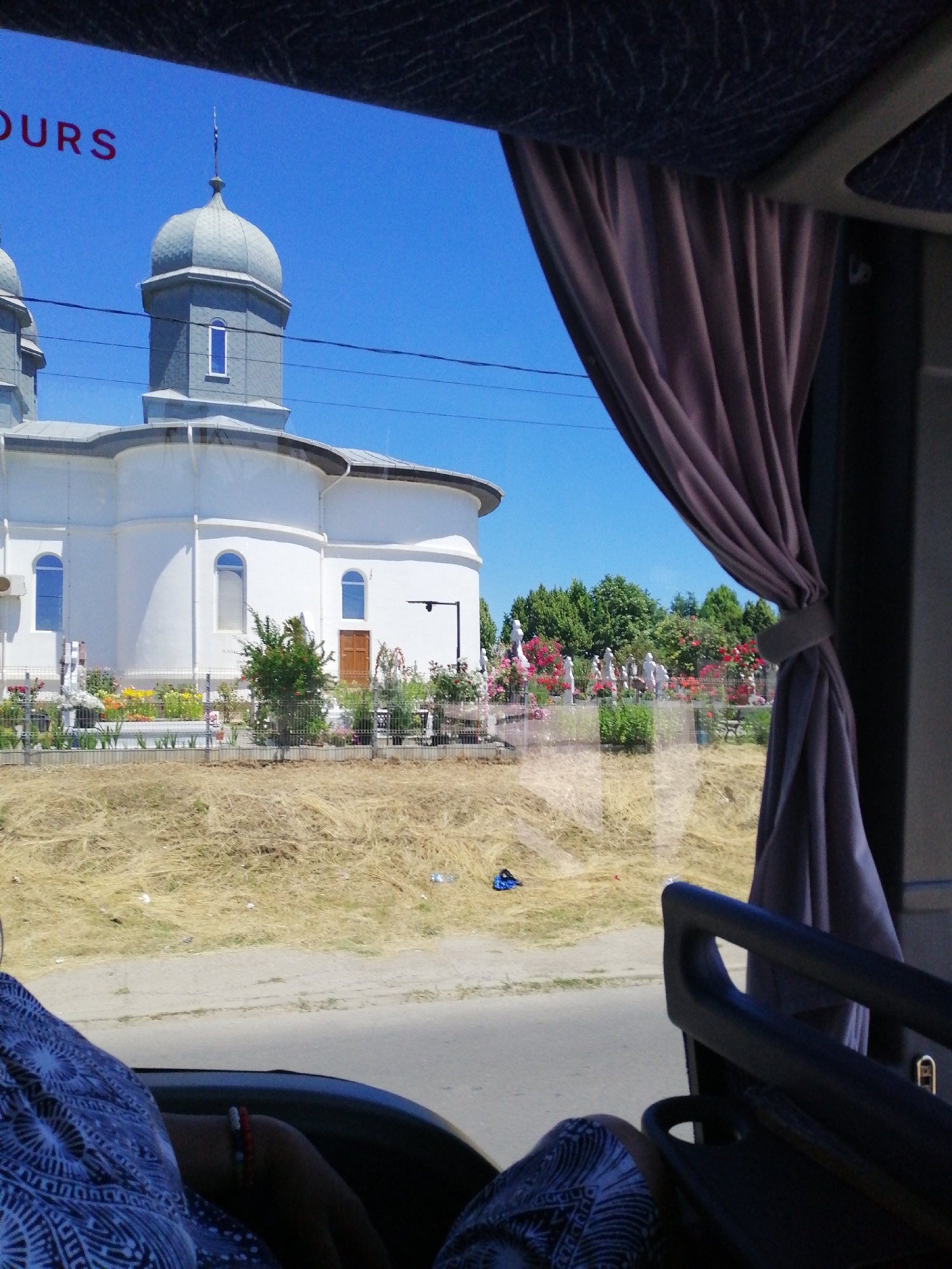 The image, taken from inside a vehicle, offers a view of a white church with grey domes against a clear blue sky. A purple curtain is partially drawn on the right side of the frame, obscuring a portion of the interior of the vehicle. In the foreground, a person's arm and shoulder are visible, wearing a patterned blue and white garment. The arm is adorned with bracelets. Outside the vehicle, a cemetery is visible, with numerous white statues and colorful flowers. The ground outside appears dry and covered in straw-like vegetation. A black fence encloses the cemetery area. The church has arched windows and a wooden door. The overall scene suggests a bright, sunny day in an outdoor setting with a religious and memorial focus.