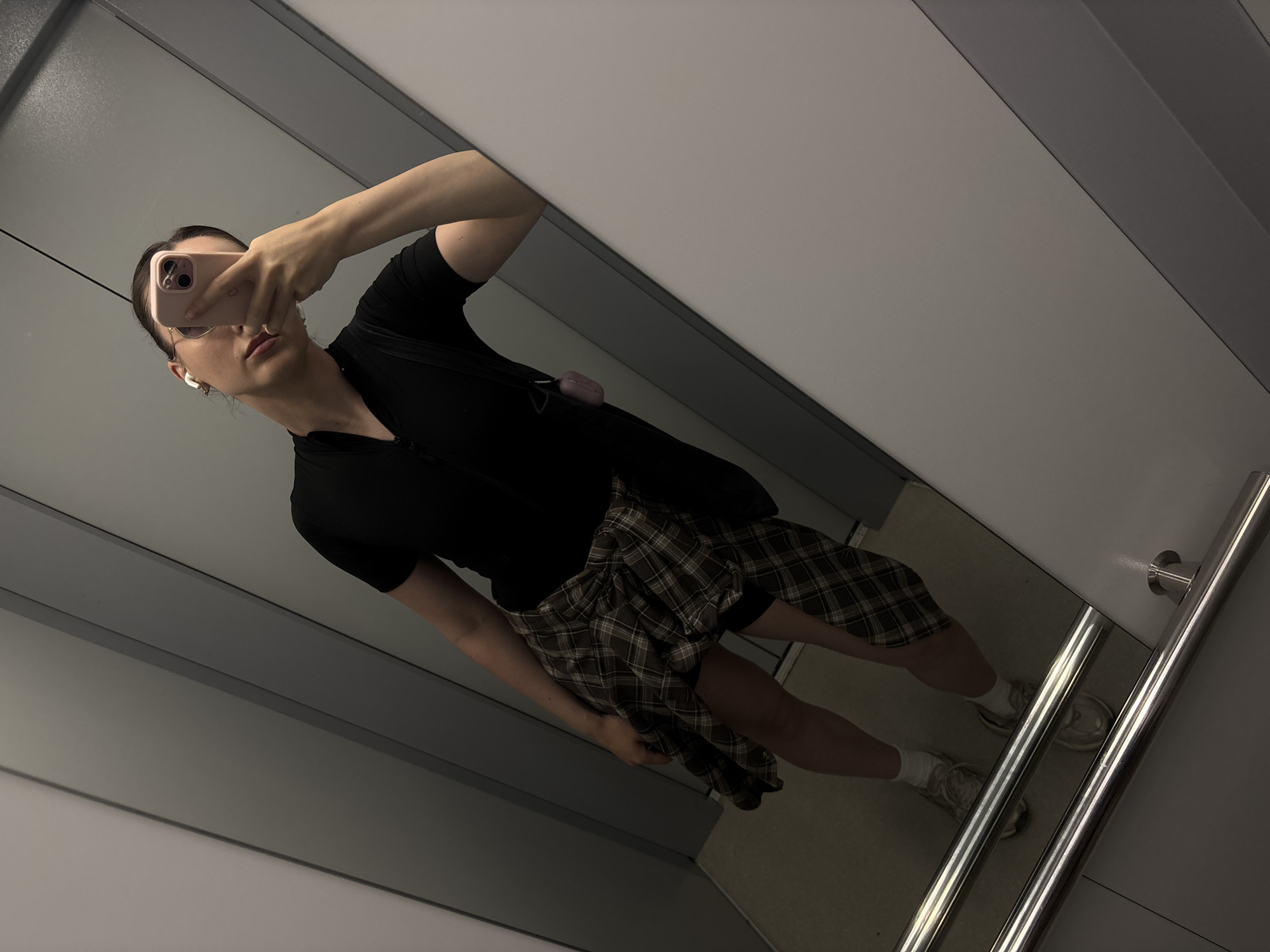 A woman is standing in an elevator, taking a mirror selfie with her phone. She is wearing a black short-sleeved top with a zipper at the neck, a plaid shirt tied around her waist, white socks, and sneakers. She has white wireless earbuds in her ears and a pink phone case. The elevator walls are grey and white, and the floor appears to be carpeted. The scene is indoors and appears to be in a modern building.