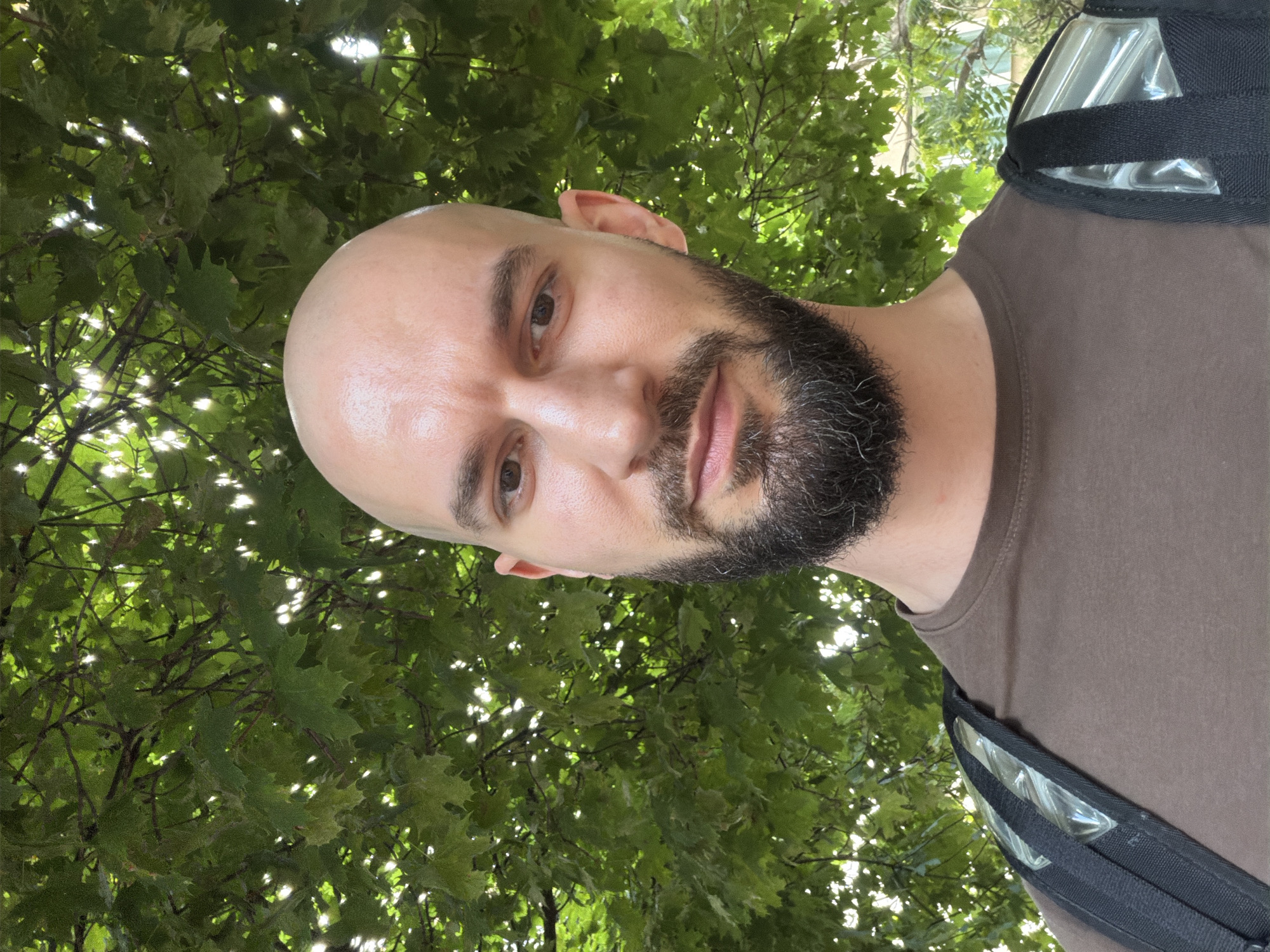 The media file is a close-up, outdoor selfie of a bald man with a beard, taken from a low angle. He is wearing a brown t-shirt and a black bag with clear compartments is visible on his shoulder. The background is dominated by lush green leaves of a tree, with sunlight filtering through, suggesting it is daytime. The man's face is the primary focus, with his eyes looking towards the camera. His expression is neutral. The overall impression is casual and relaxed, possibly taken during a walk or outdoor activity. There is no visible text in the media file. The location context is Sofia, Bulgaria.