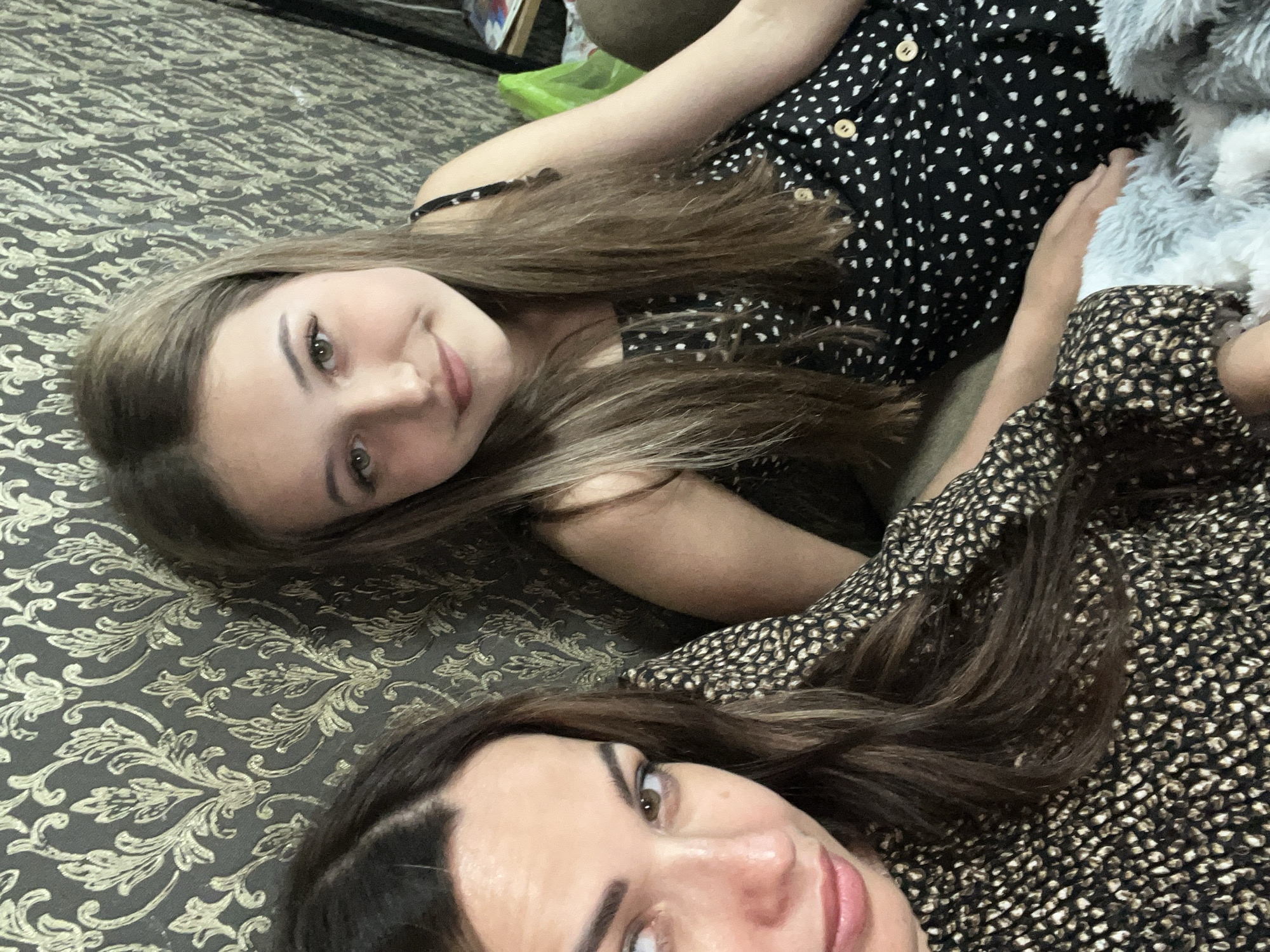 In this medium shot, two females are seated on a couch with a patterned wallpapered background. The female on the left, in the foreground, has dark brown hair and is wearing a black dress with a speckled gold and black pattern. She is looking to her left with her mouth slightly open. The female on the right, slightly behind and to the right of the first, has long, light brown hair and is wearing a black dress with white polka dots. She is looking forward and smiling. A fluffy grey blanket is visible in the bottom right corner of the image. The overall lighting is soft and natural. The background features a damask-style wallpaper with a metallic gold pattern on a dark grey base.